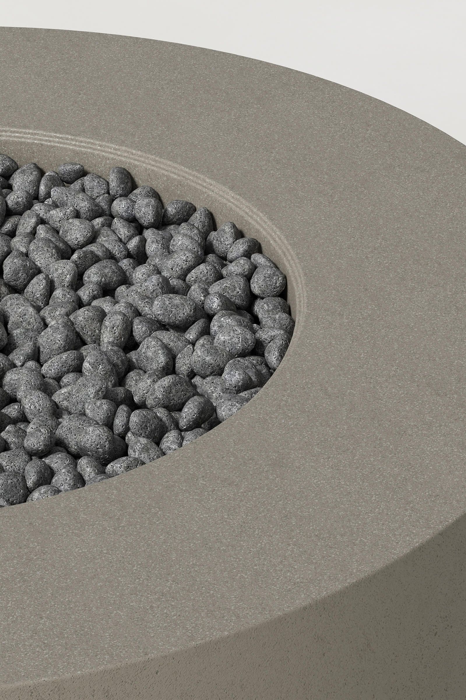 Close-up photograph of a cylindrical dark grey concrete fire pit with thick walls and a well filled with volcanic rocks. Showcases a smooth texture and precise edge design.