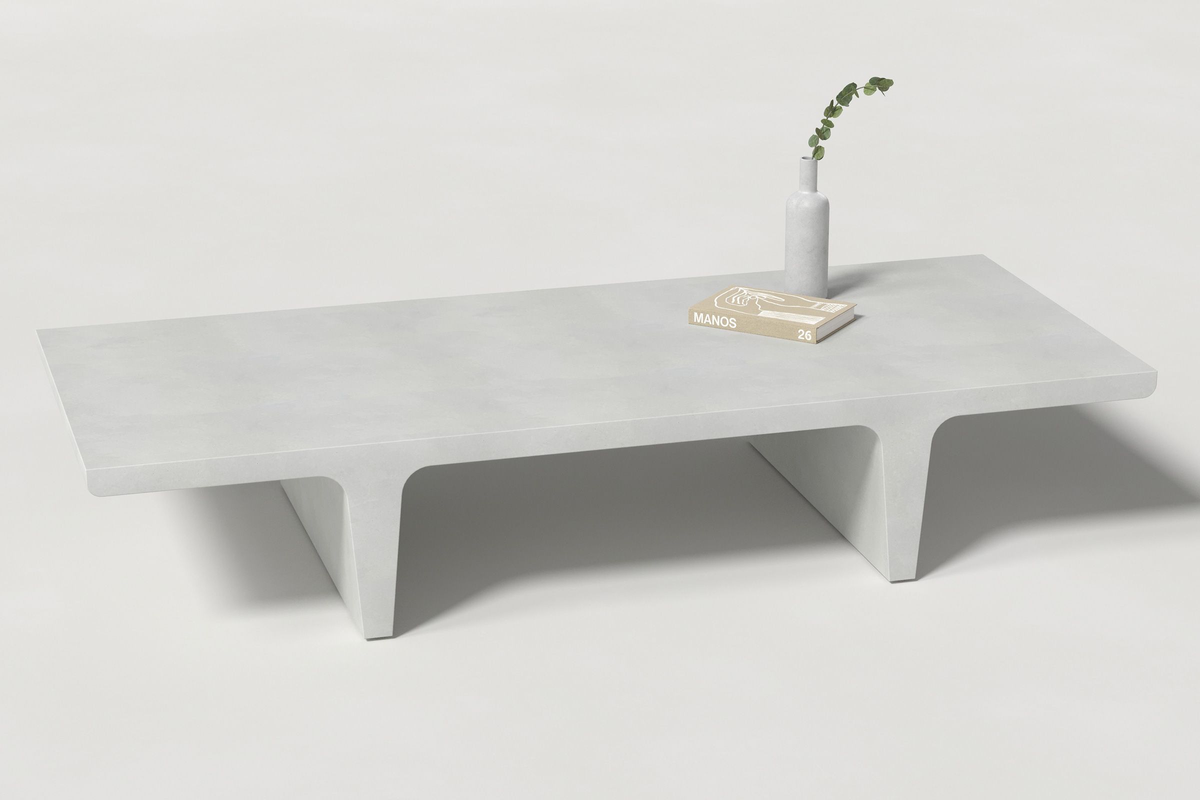A rectangular white concrete coffee table with sculptural double tee legs, styled with a neutral-toned book and a slender vase holding a eucalyptus, set against a clean white background.