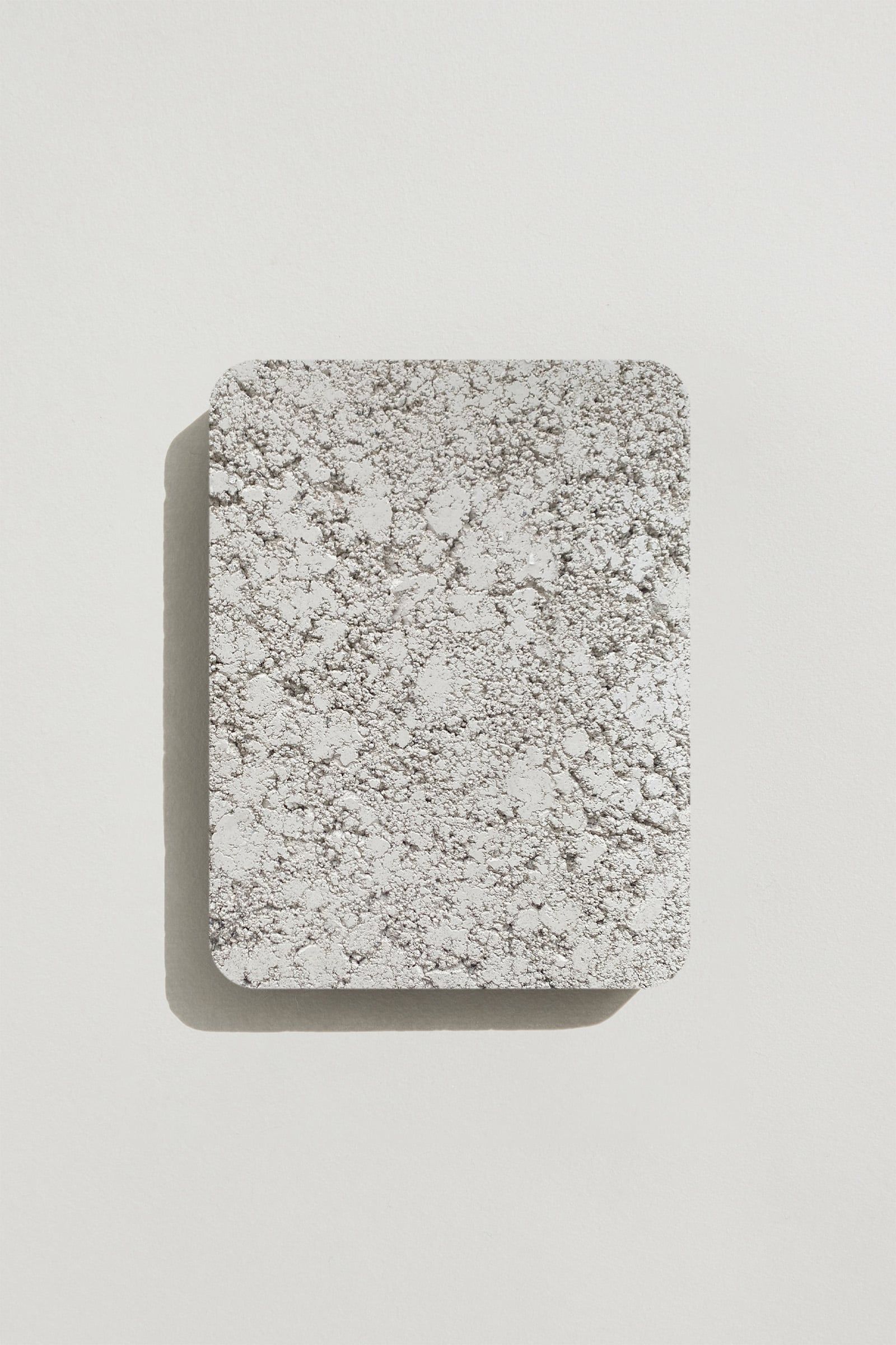 A rectangular sample of rough white concrete with a natural, pitted surface texture. Photographed on a white background with a cast shadow.