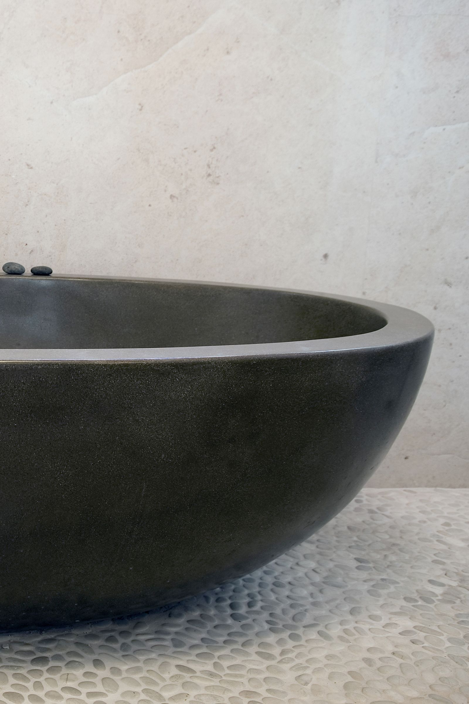 A close-up view of a smooth, dark concrete bathtub with rounded edges, set against a light stone wall. Three small black stones rest on the tub’s edge, and the floor is covered in textured pebble-like tiles.