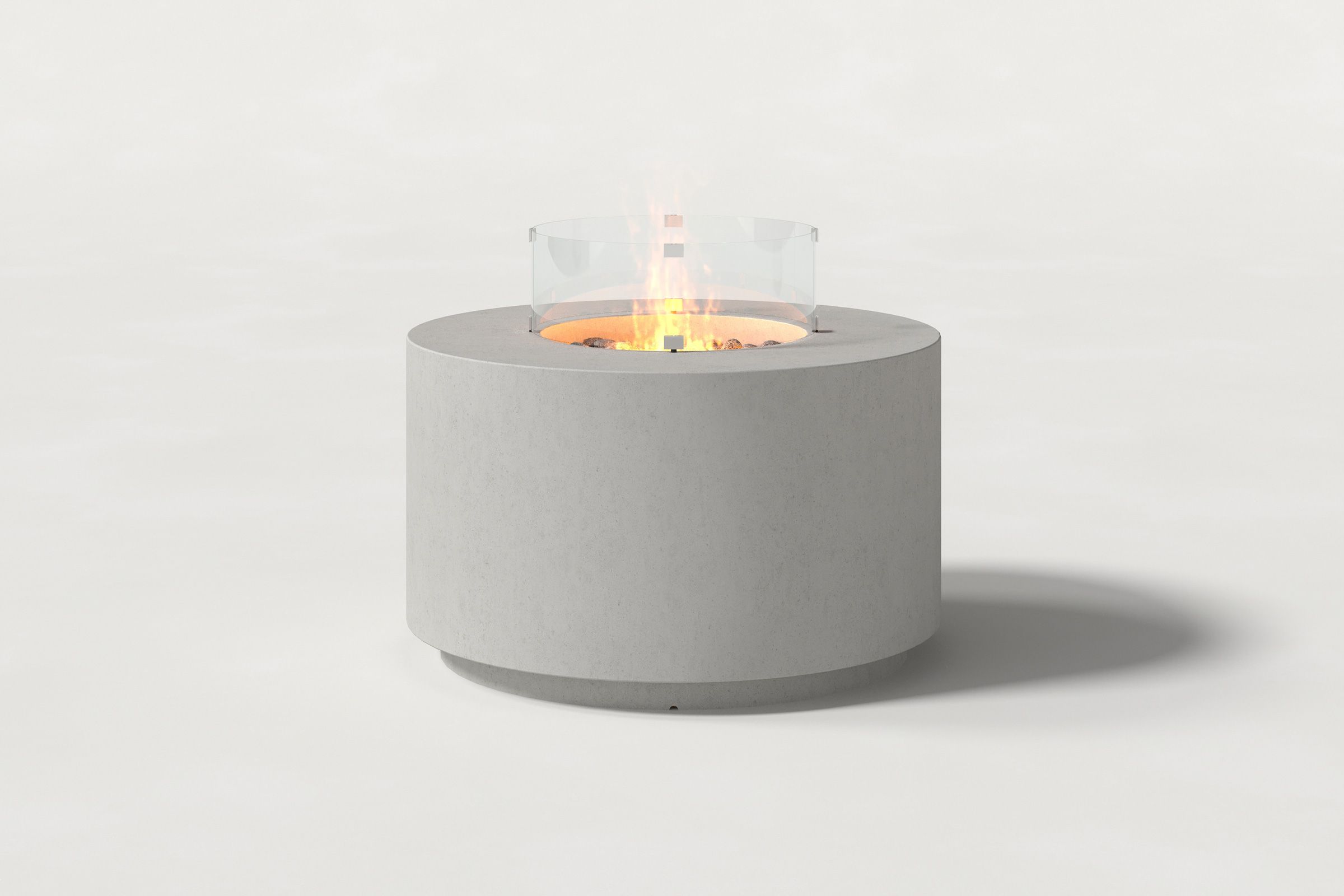 Low cylindrical light grey colored concrete fire pit with thick walls and a central circular well filled with dark volcanic rocks and flame, surrounded by a clear glass shield. Side view on a white background with a shadow to the right.