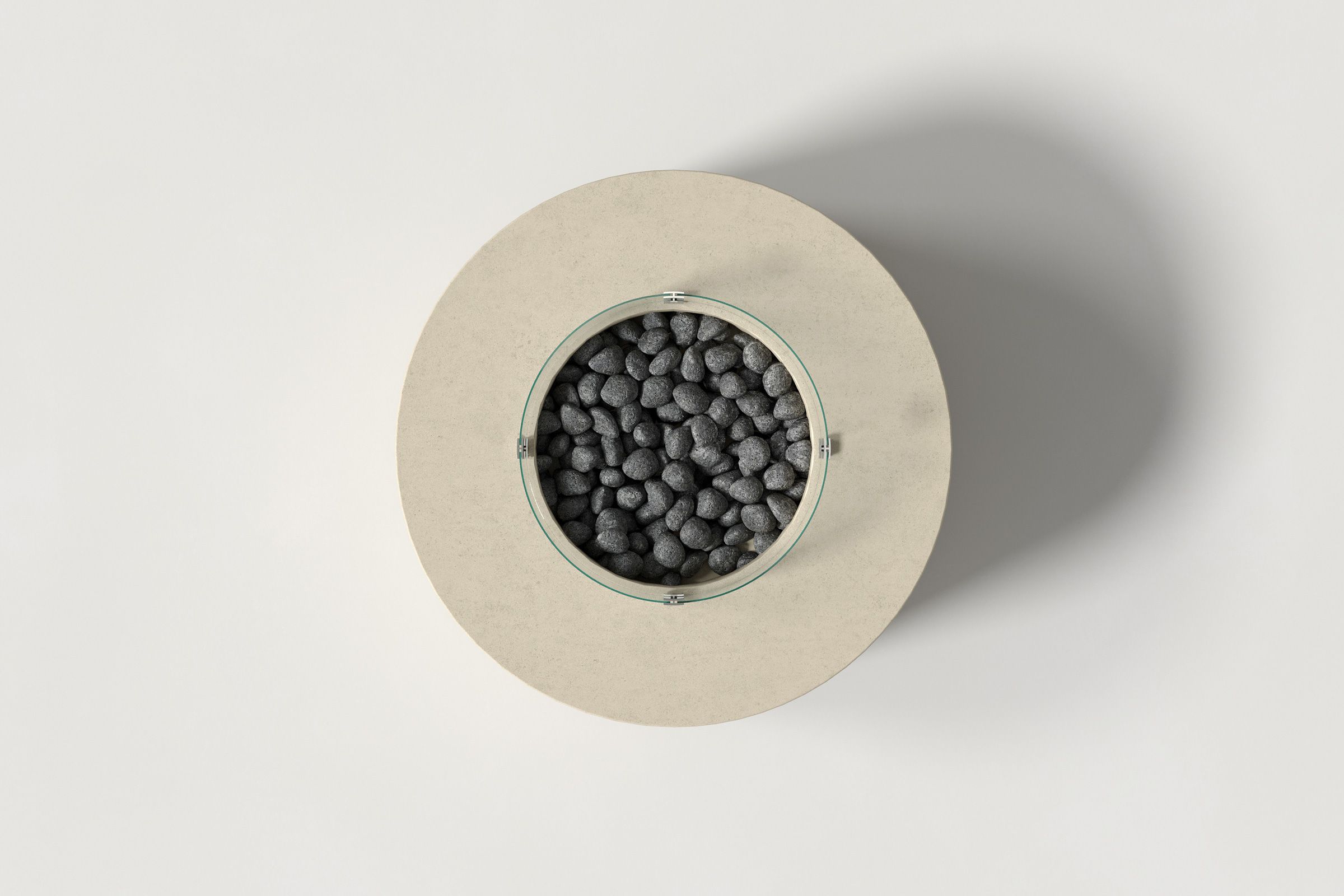 Top-down view of a circular sand-colored concrete fire pit with thick walls and a central well filled with dark volcanic rocks, encircled by a clear glass shield on a white background.