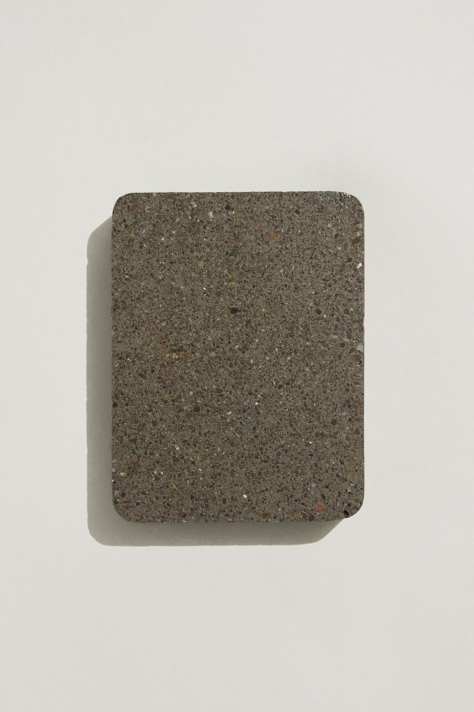 A rectangular sample of dark grey concrete with small black, grey and brown aggregate speckles, and rounded edges. Photographed on a white background with a cast shadow