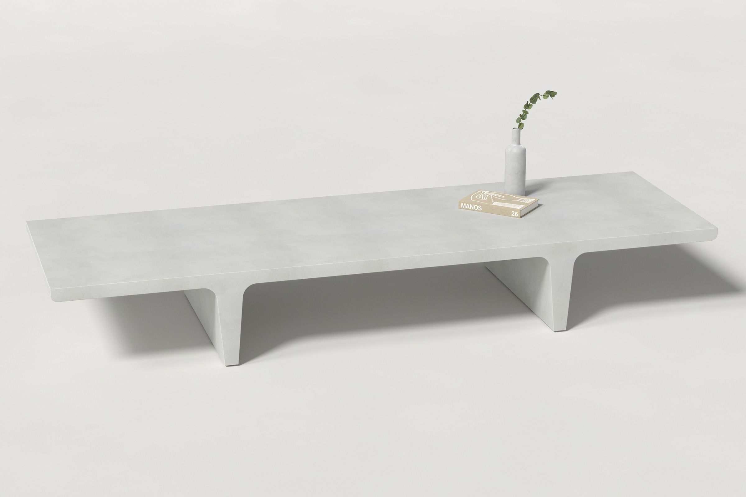 A long rectangular white concrete coffee table with sculptural double tee legs, styled with a neutral-toned book and a slender vase holding a eucalyptus, set against a clean white background.