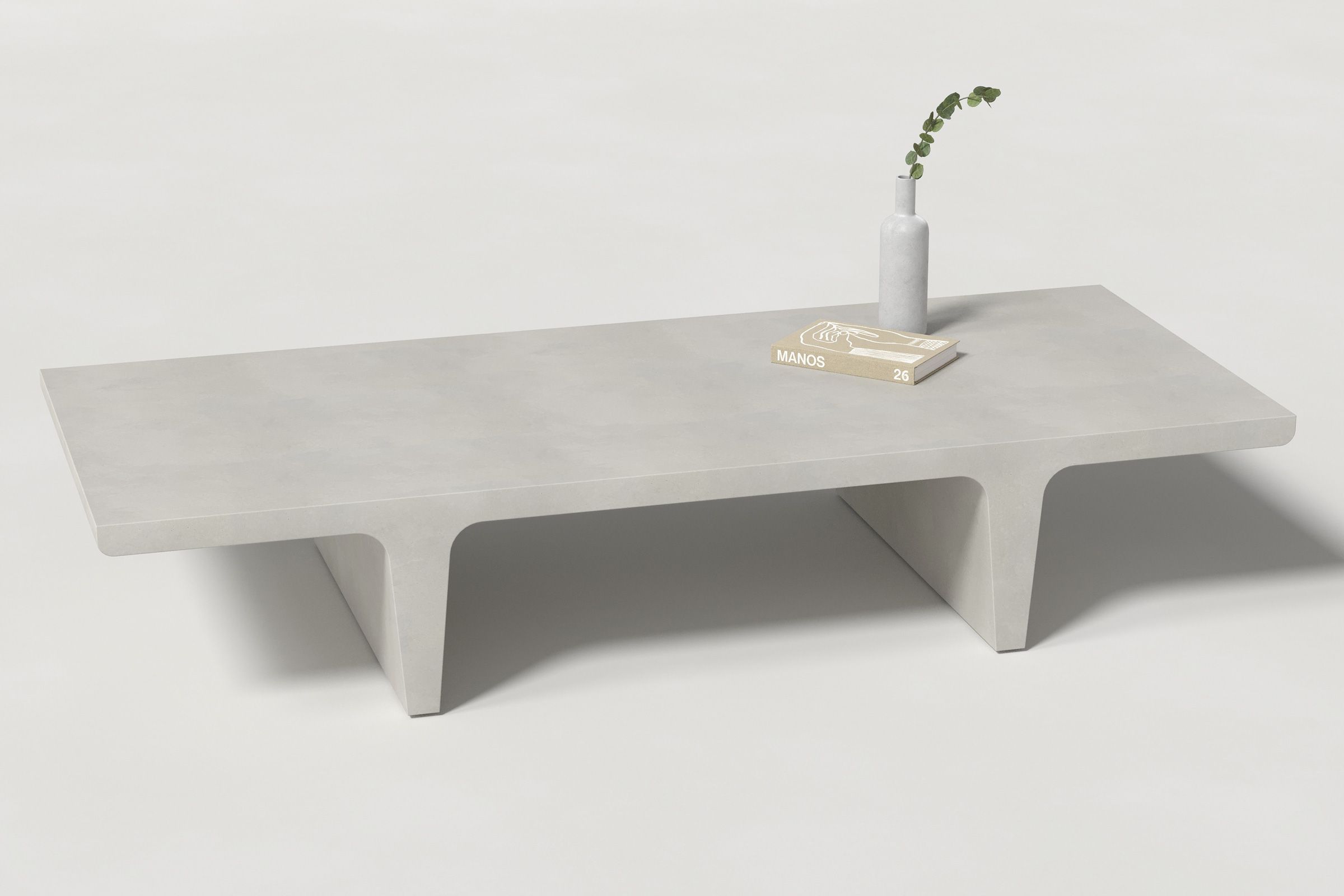 A rectangular light grey concrete coffee table with sculptural double tee legs, styled with a neutral-toned book and a slender vase holding a eucalyptus, set against a clean white background.