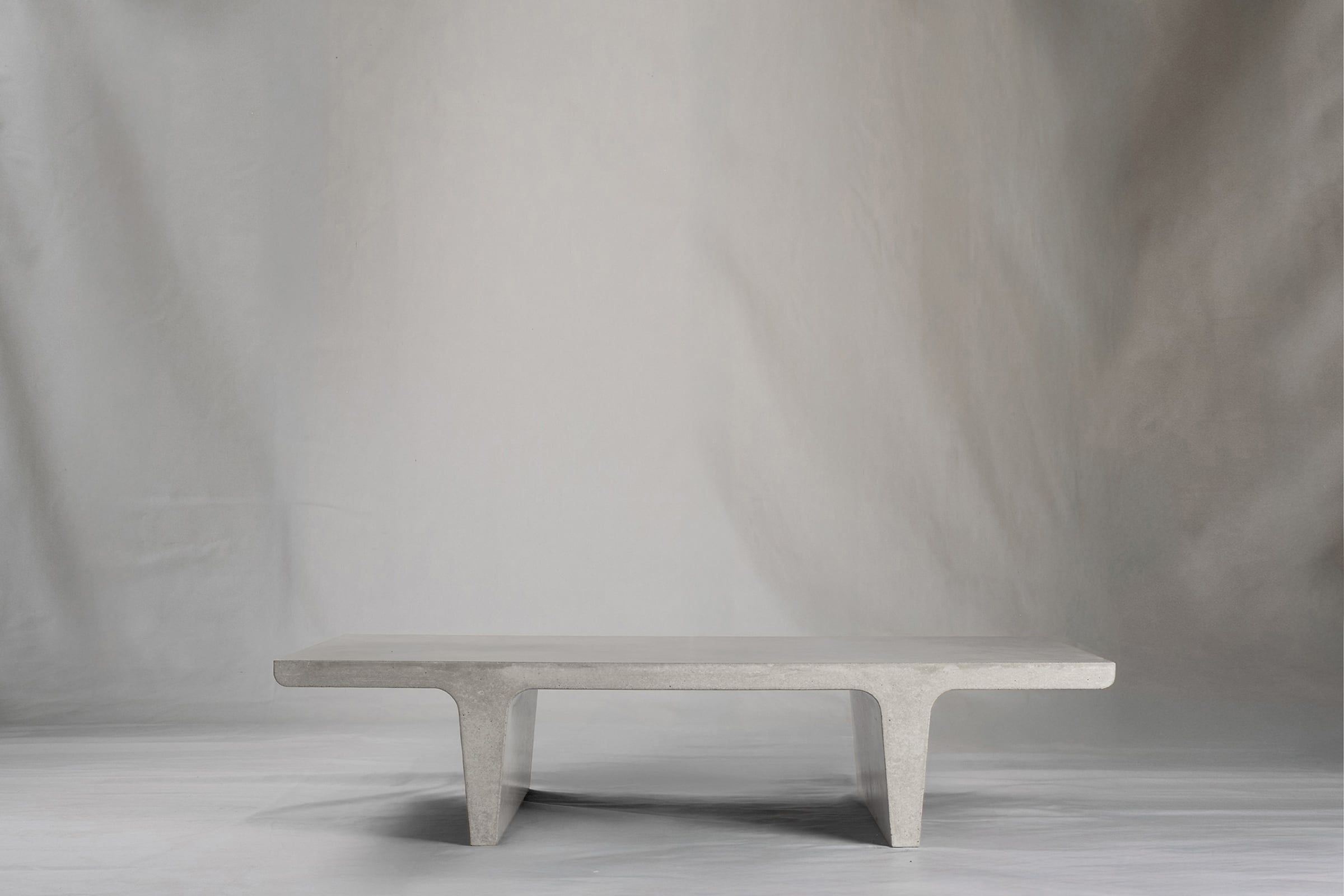 Rectangular light grey concrete coffee table with sculptural double tee legs, featuring a smooth troweled finish, set against a soft neutral backdrop.