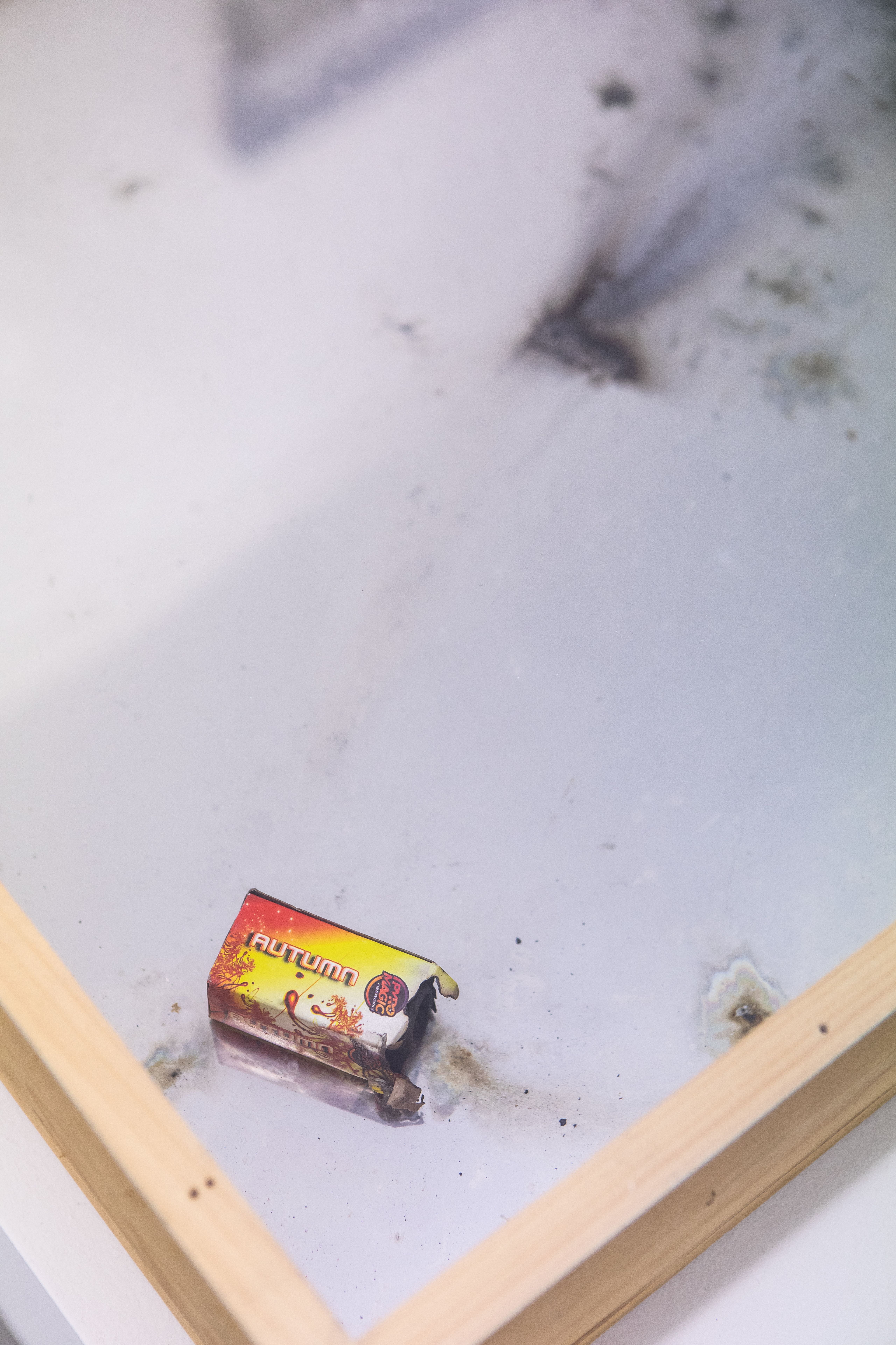 Exploded firecracker box inside a wooden frame, with soot on the glass