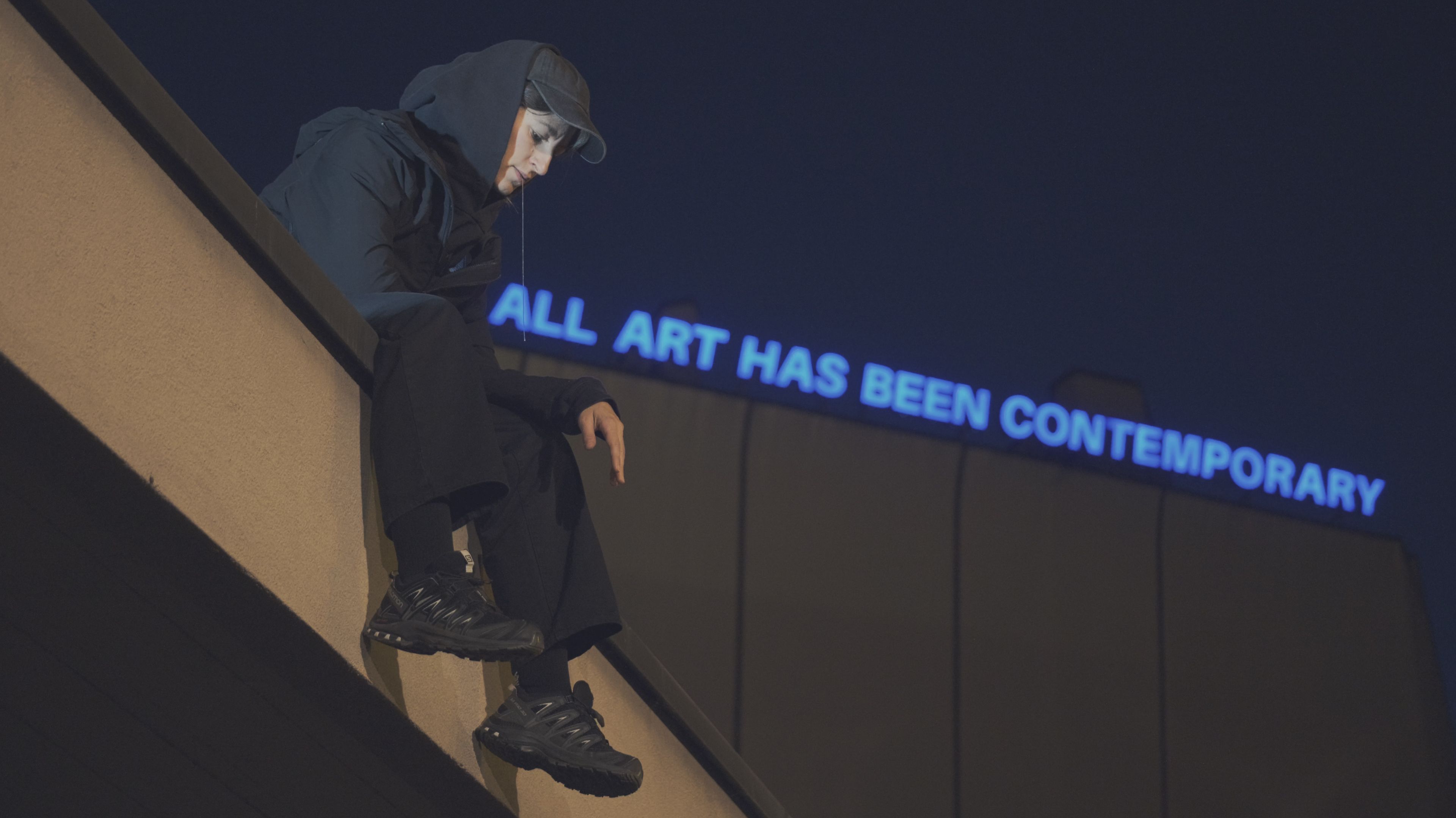 Hooded person sitting on a sloping wall beneath a neon sign reading ALL ART HAS BEEN CONTEMPORARY