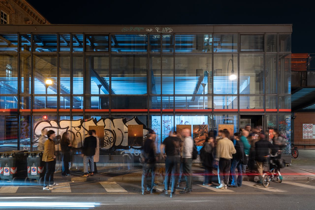 Glass building at night lit blue inside, with visitors gathered on the pavement in front