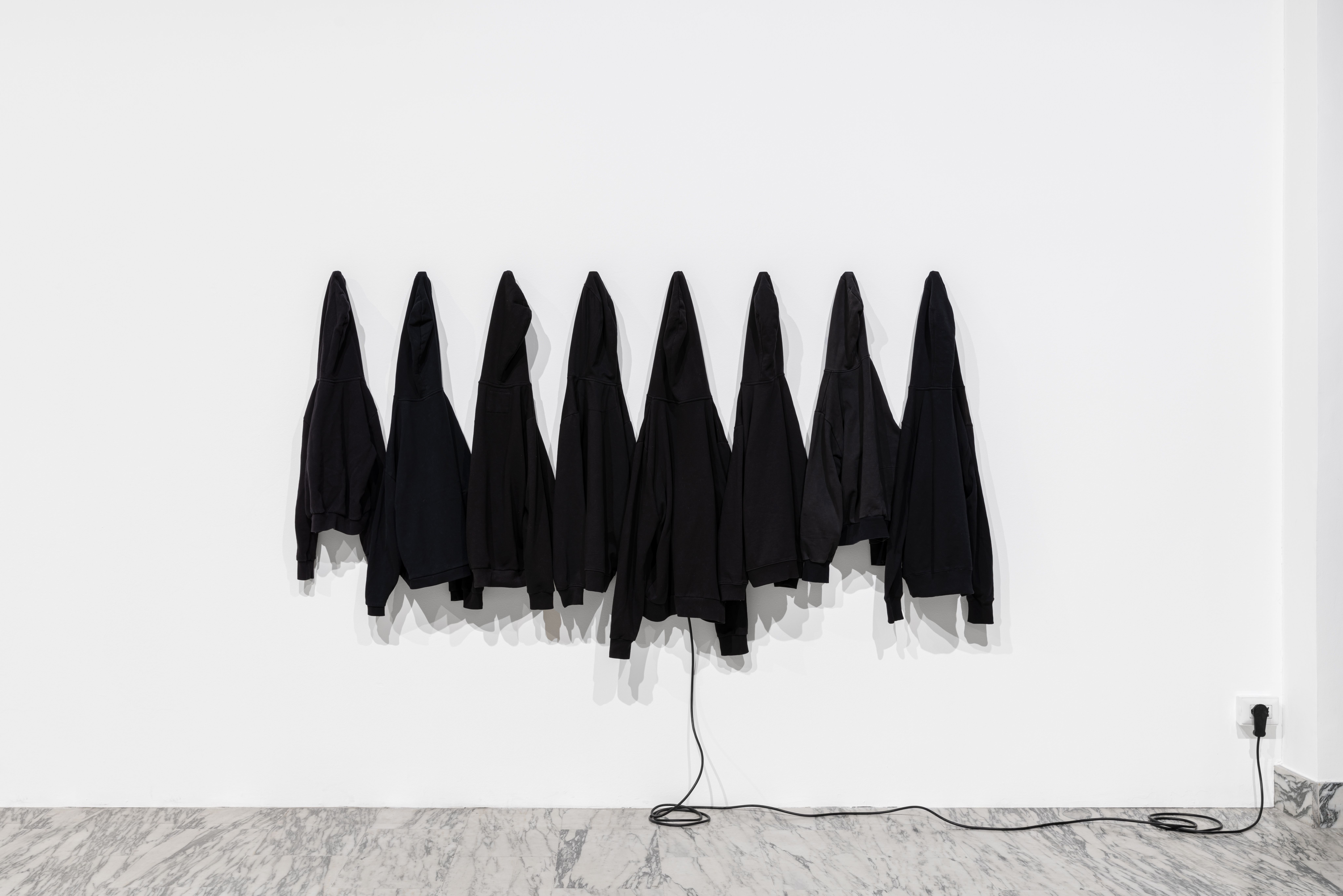 Eight black sweatshirts hung in a row on a white wall, a cable running to a socket