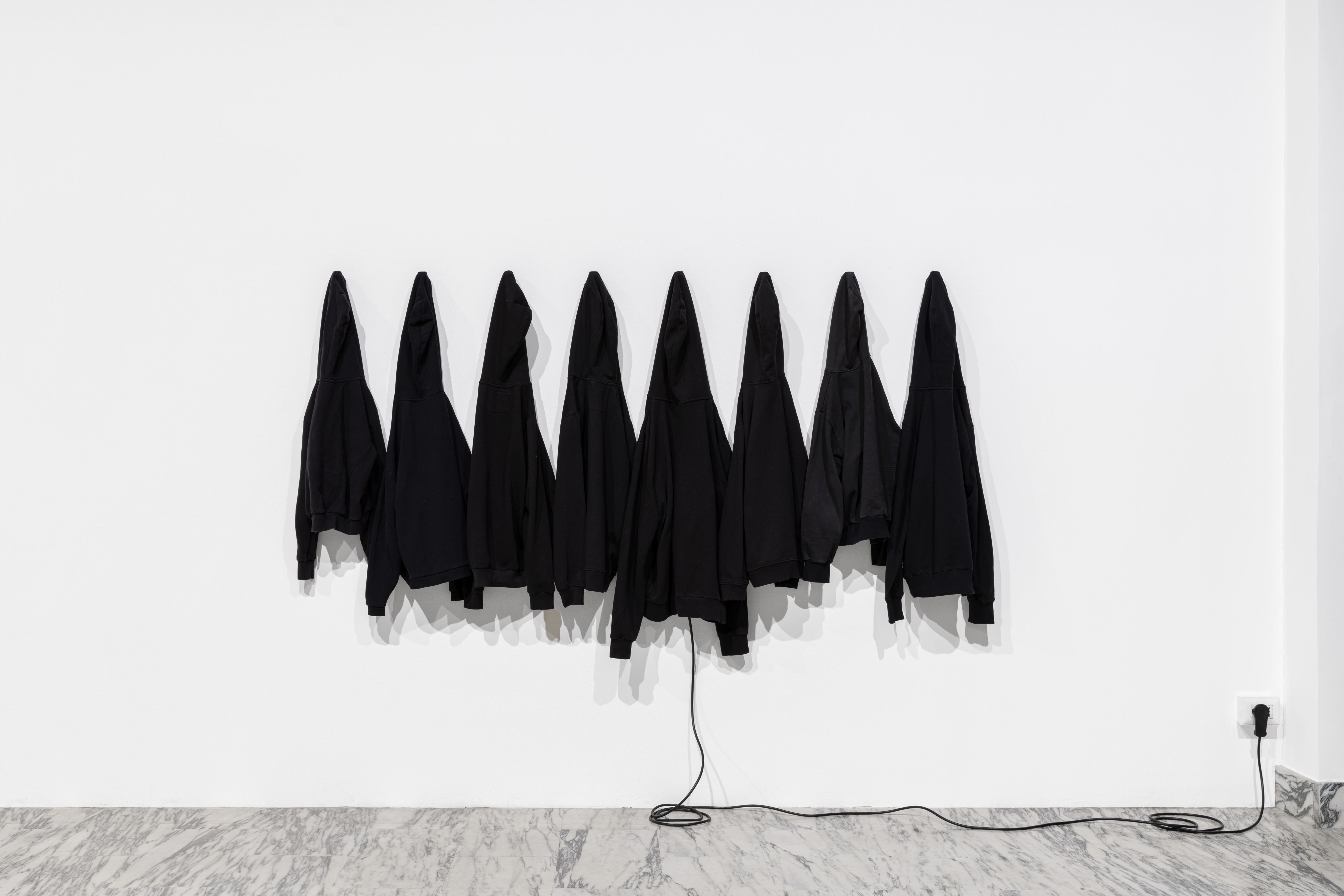 Eight black sweatshirts hung in a row on a white wall, a cable running to a socket