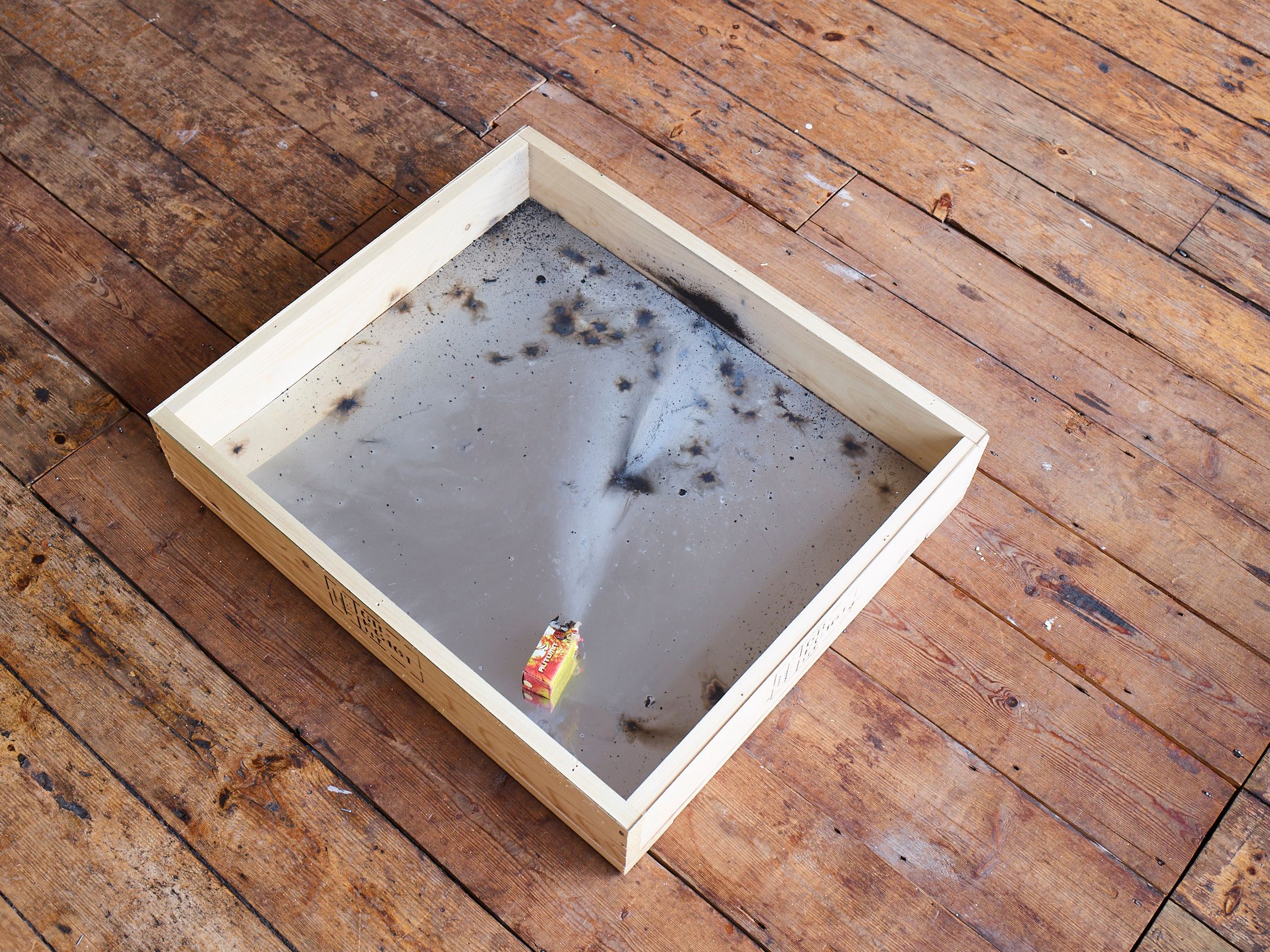 Wooden case on a plank floor, with soot and a spent firecracker under glass
