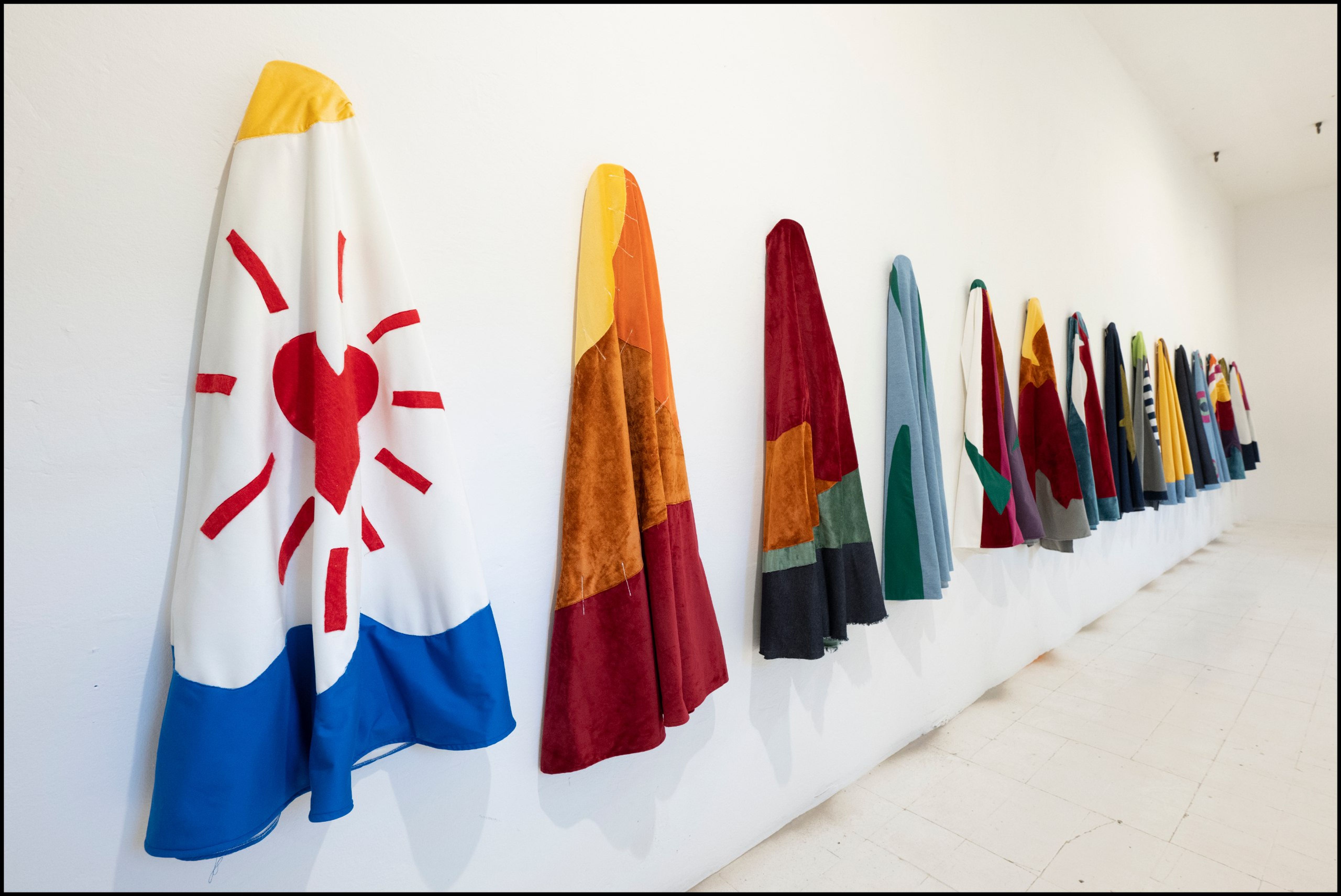 Capes in patchwork fabrics hanging in a long row on a white wall, the first with a red heart and sun