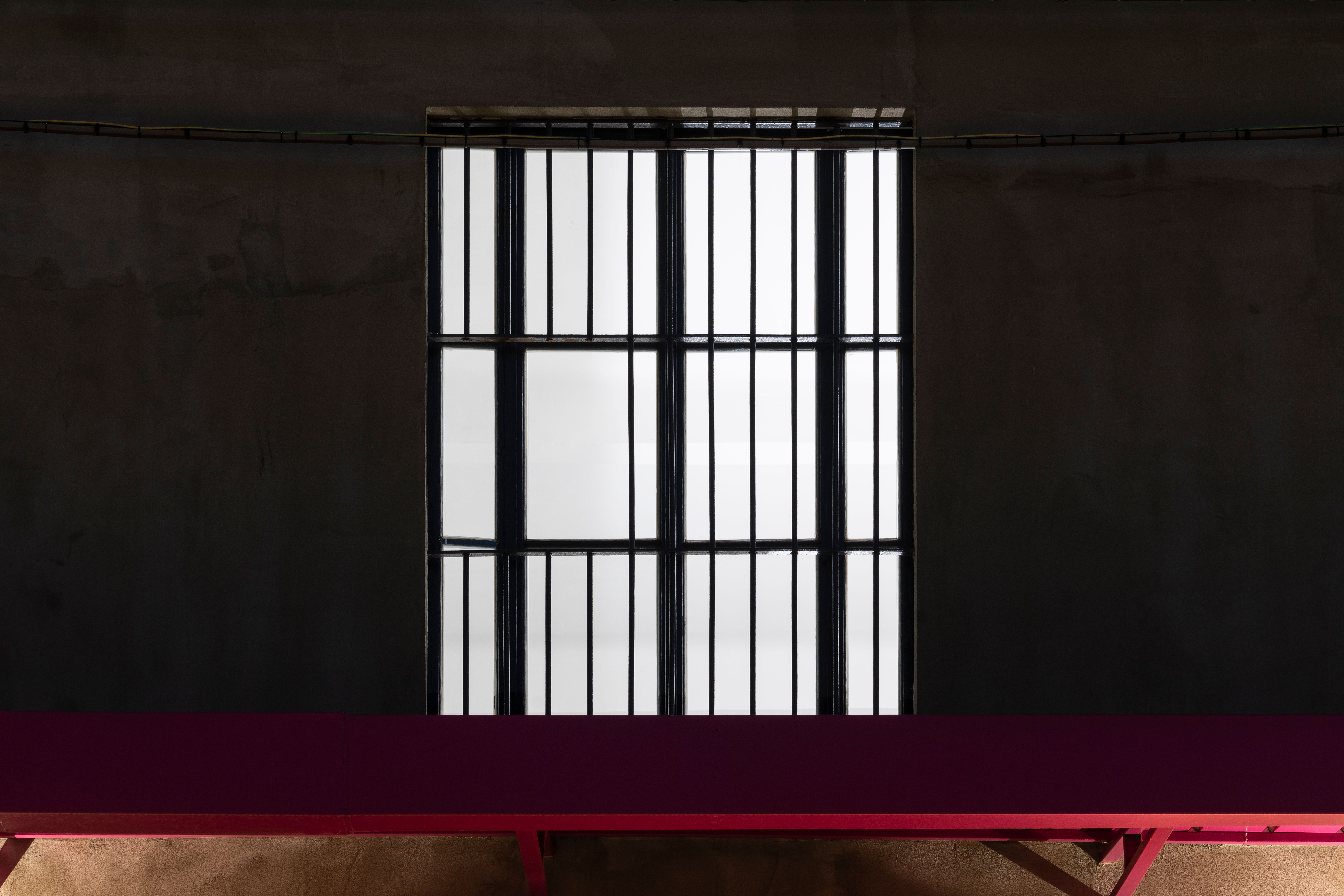 Large barred window glowing white on a dark wall above a red bench