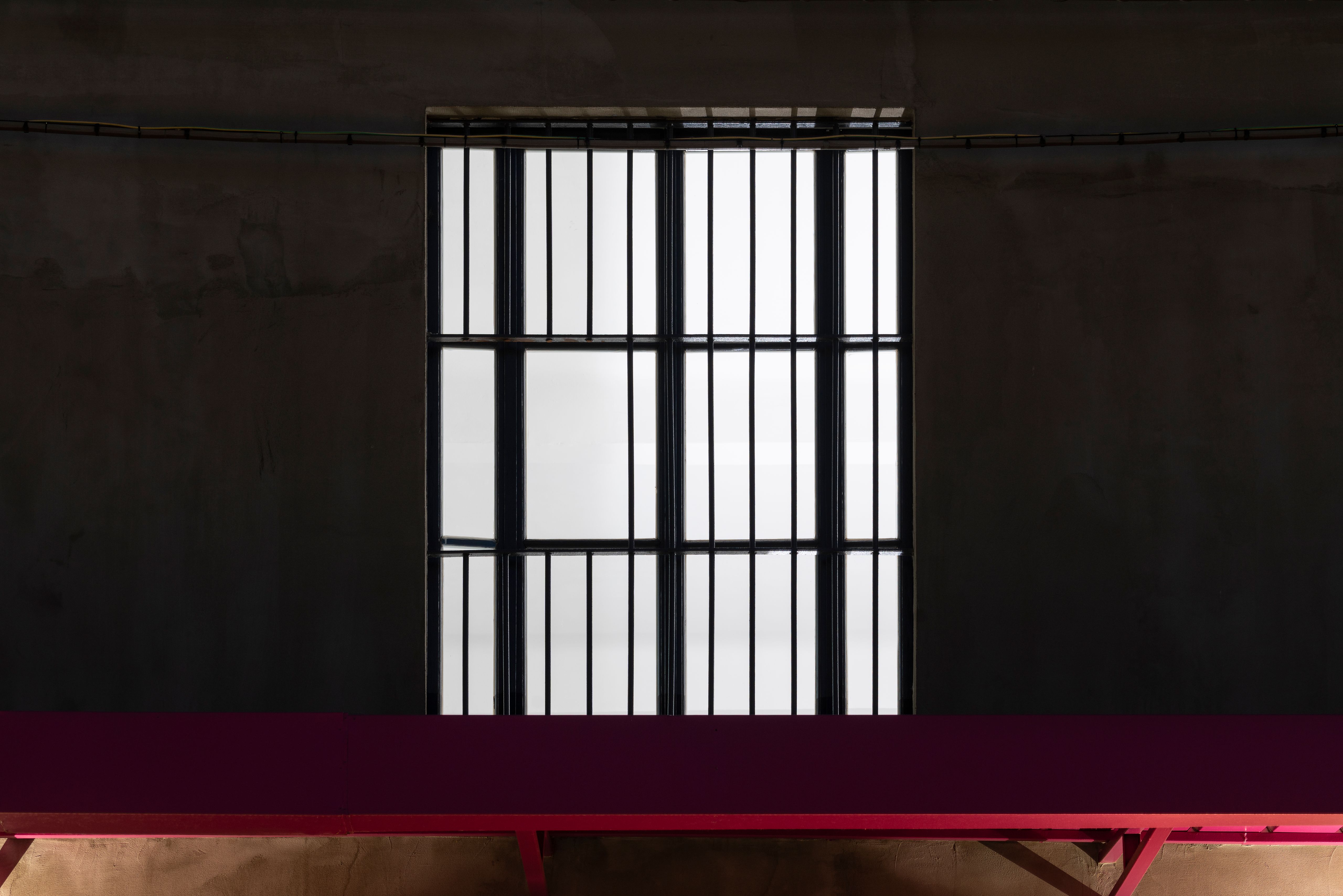 Large barred window glowing white on a dark wall above a red bench