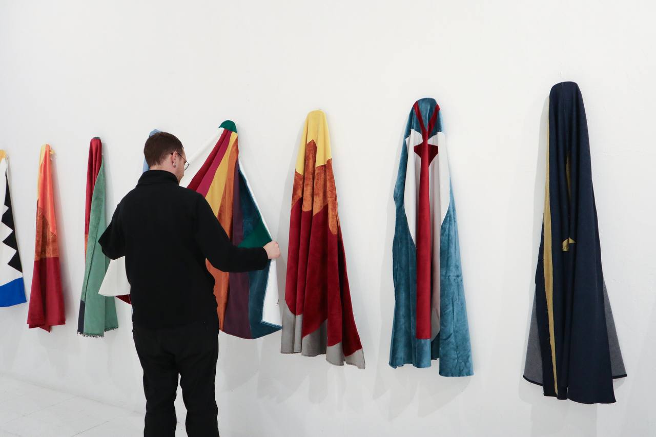 A visitor handles one of the capes hanging in a row on a white wall