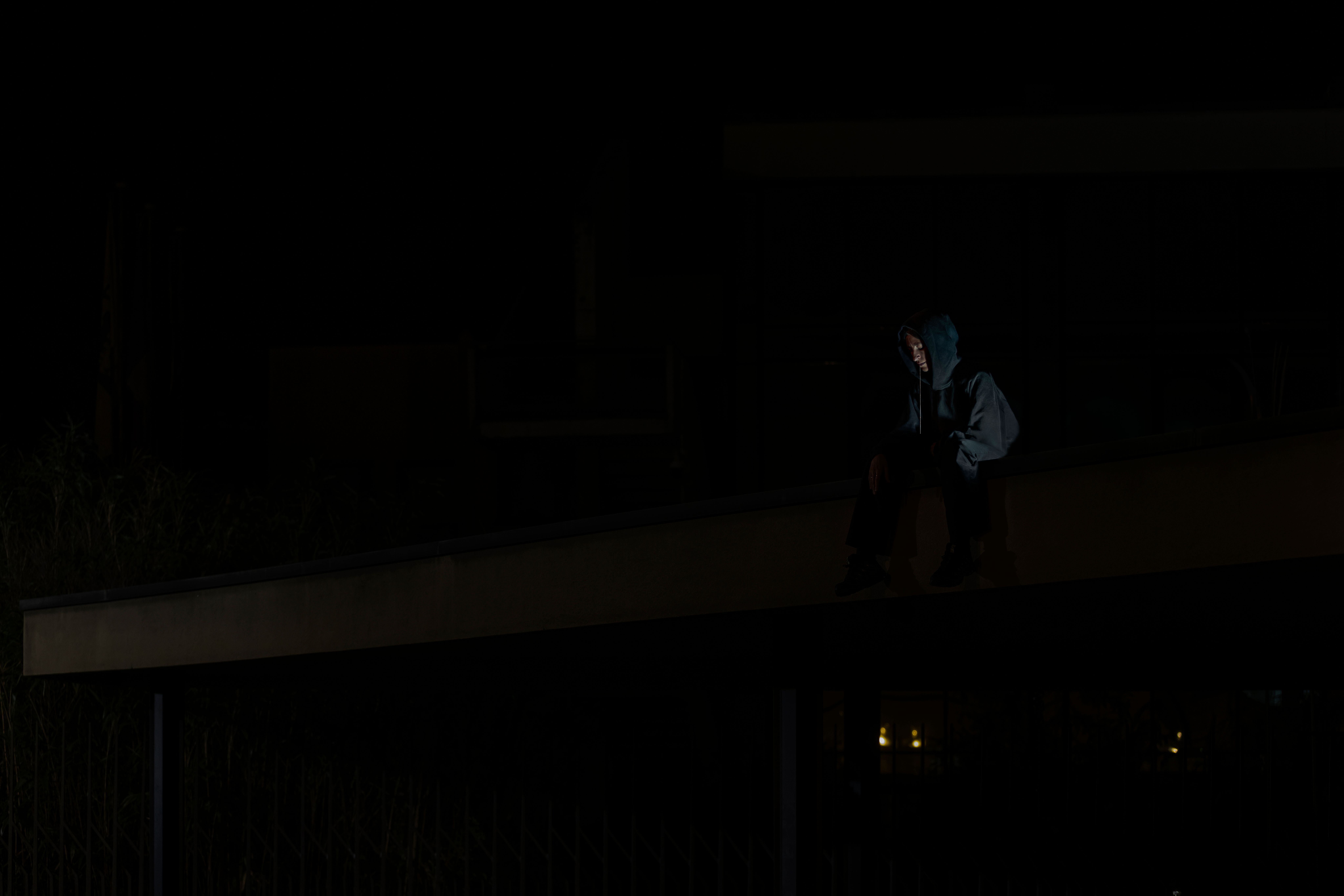 Figure in a hooded jacket sitting on a ledge among dark trees and buildings
