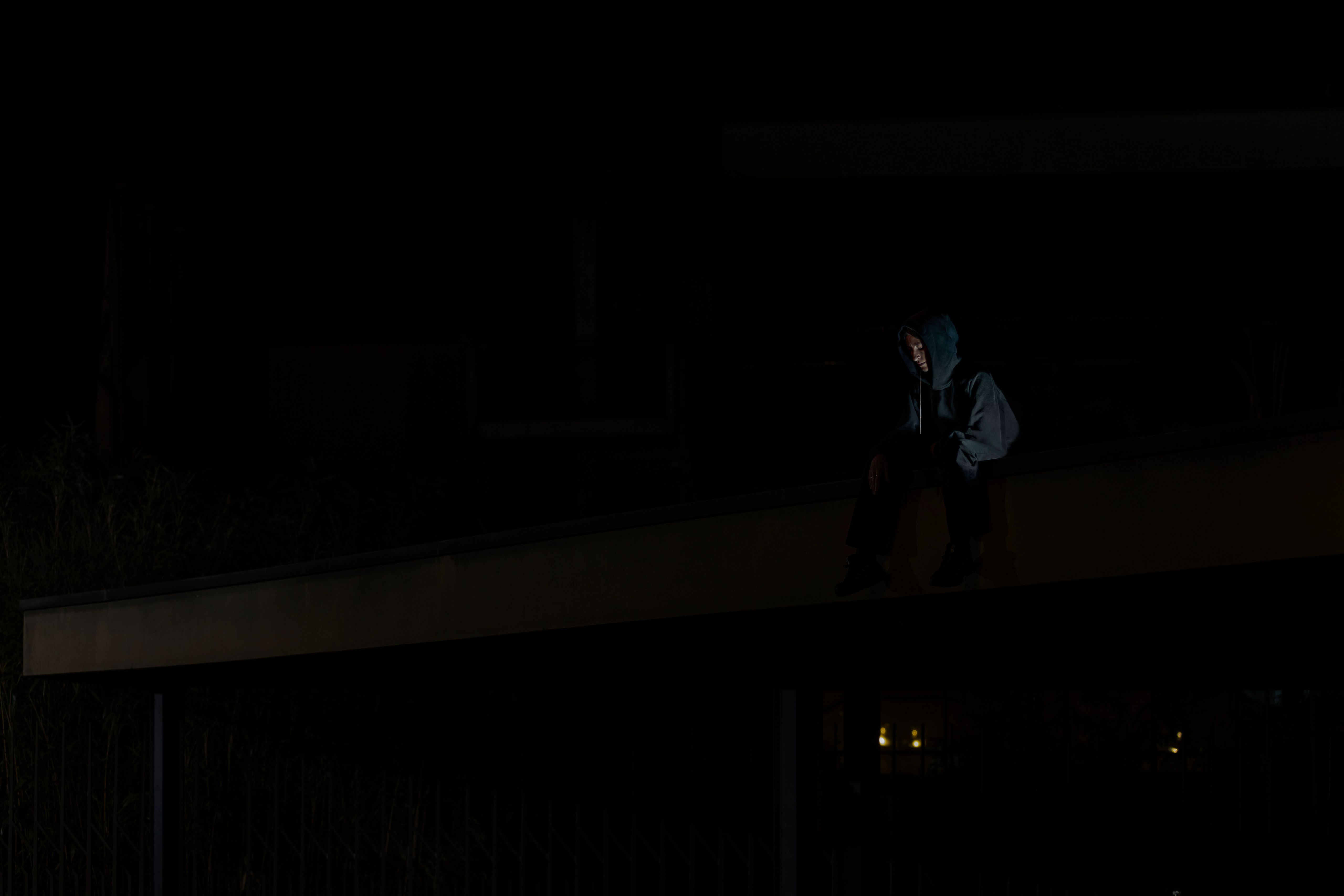 Figure in a hooded jacket sitting on a ledge among dark trees and buildings