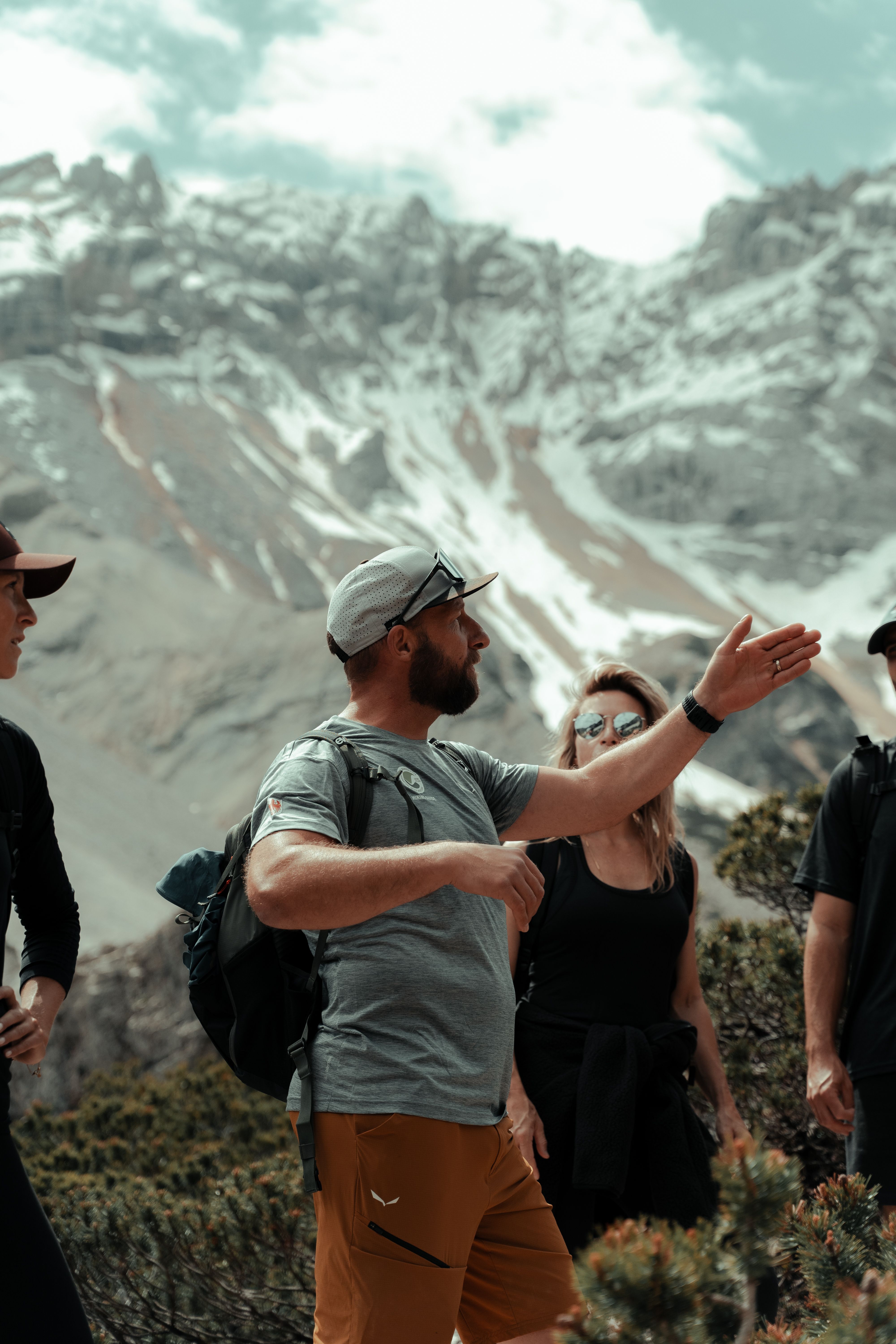 Group of hikers standing outdoors in a mountainous area, with a bearded man in the foreground gesturing as if explaining or pointing something out, and snow-capped peaks visible in the background.