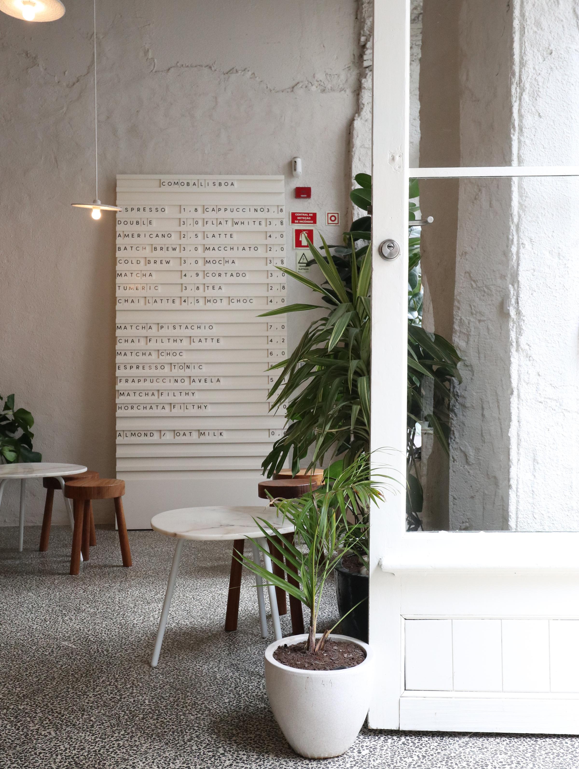 A modern, minimalist coffee shop interior featuring a light and airy design. The space includes a terrazzo floor, small white and wooden tables with matching stools, potted plants, and a menu board displayed on the wall with various coffee and drink options. A white-framed glass door partially opens into the area, adding to the clean aesthetic.