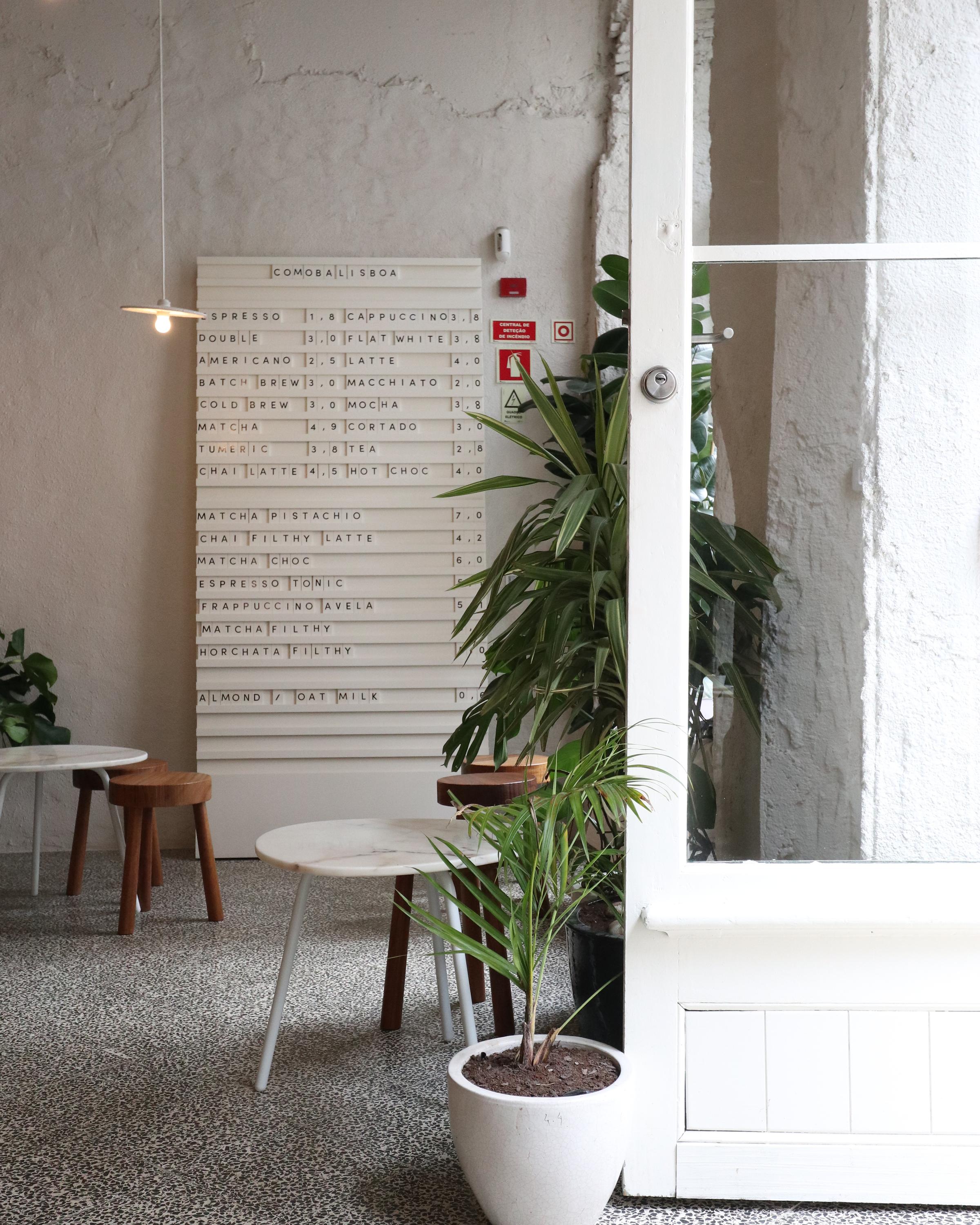 A modern, minimalist coffee shop interior featuring a light and airy design. The space includes a terrazzo floor, small white and wooden tables with matching stools, potted plants, and a menu board displayed on the wall with various coffee and drink options. A white-framed glass door partially opens into the area, adding to the clean aesthetic.
