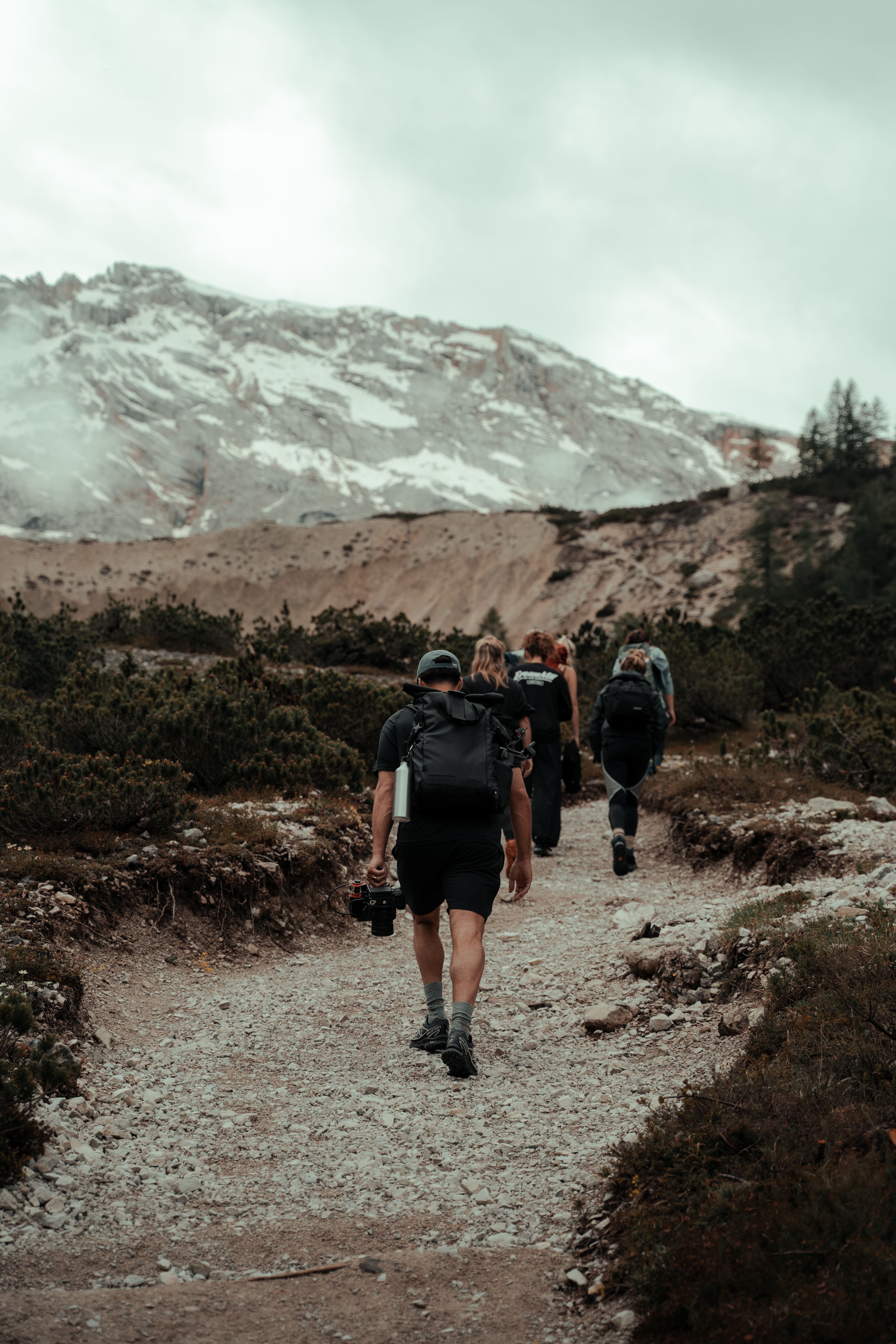 A group of hikers walking along a rocky trail in a mountainous landscape, with snow-capped peaks and cloudy skies in the background, surrounded by sparse vegetation and rugged terrain.