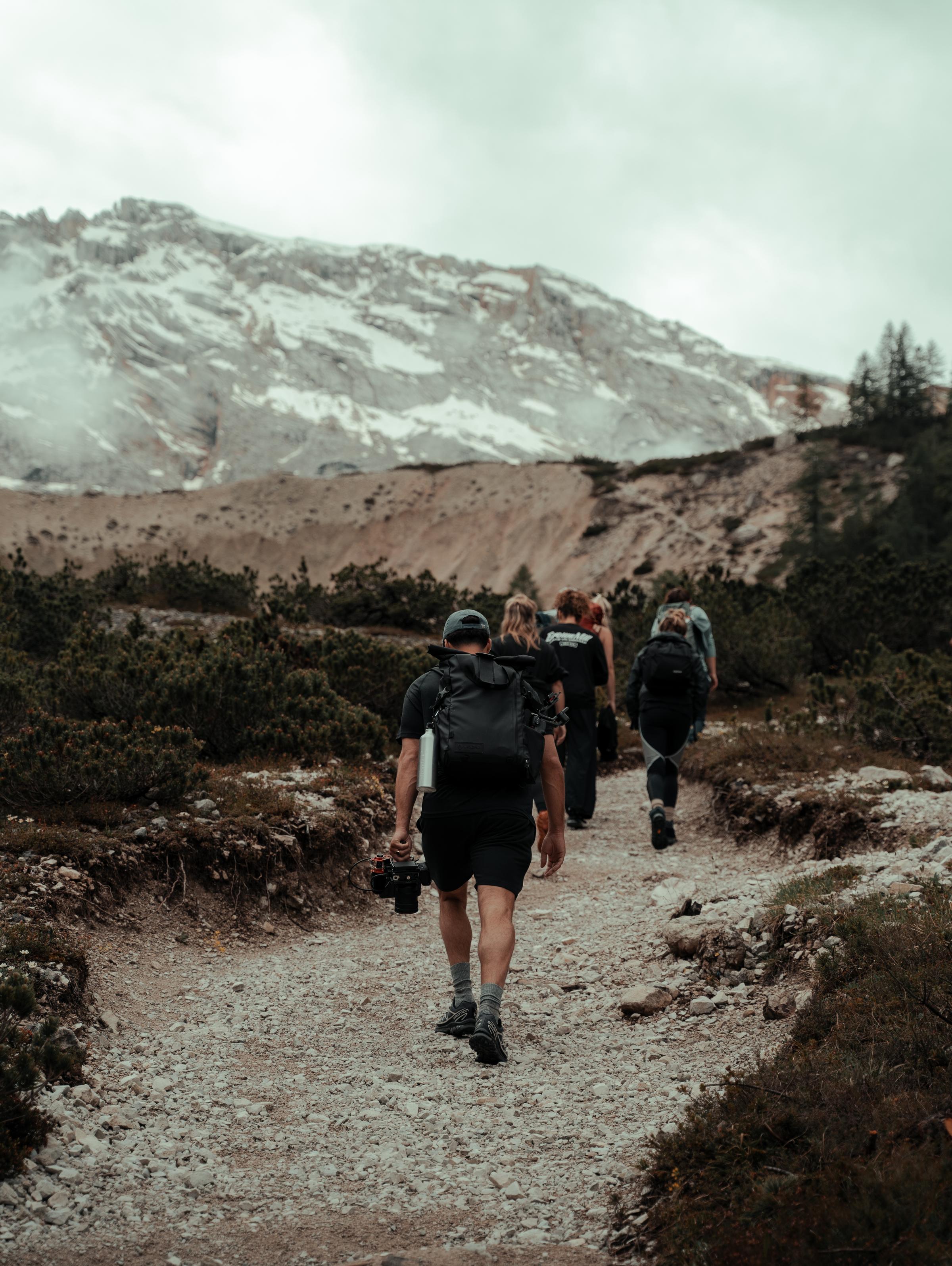 A group of hikers walking along a rocky trail in a mountainous landscape, with snow-capped peaks and cloudy skies in the background, surrounded by sparse vegetation and rugged terrain.