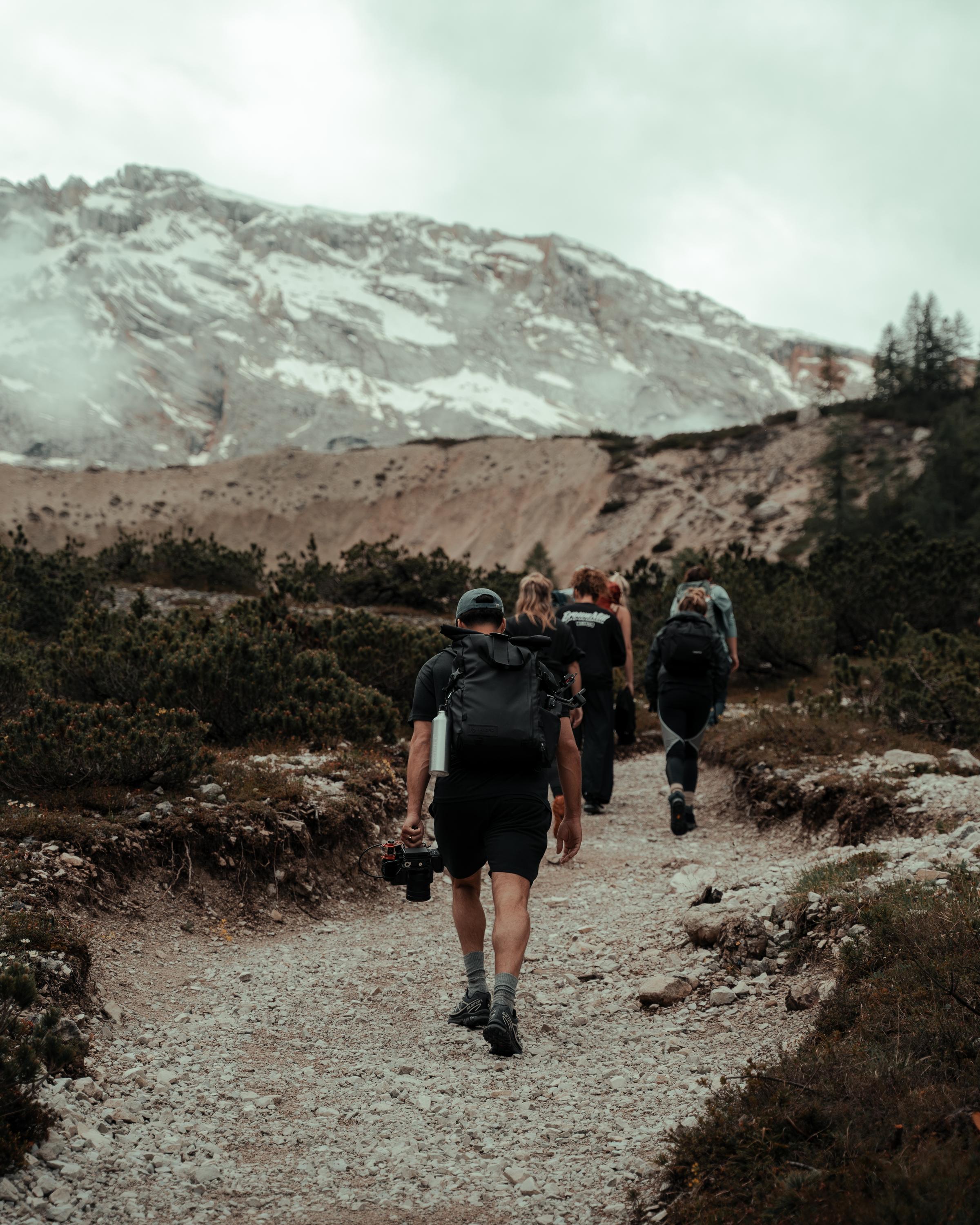 A group of hikers walking along a rocky trail in a mountainous landscape, with snow-capped peaks and cloudy skies in the background, surrounded by sparse vegetation and rugged terrain.