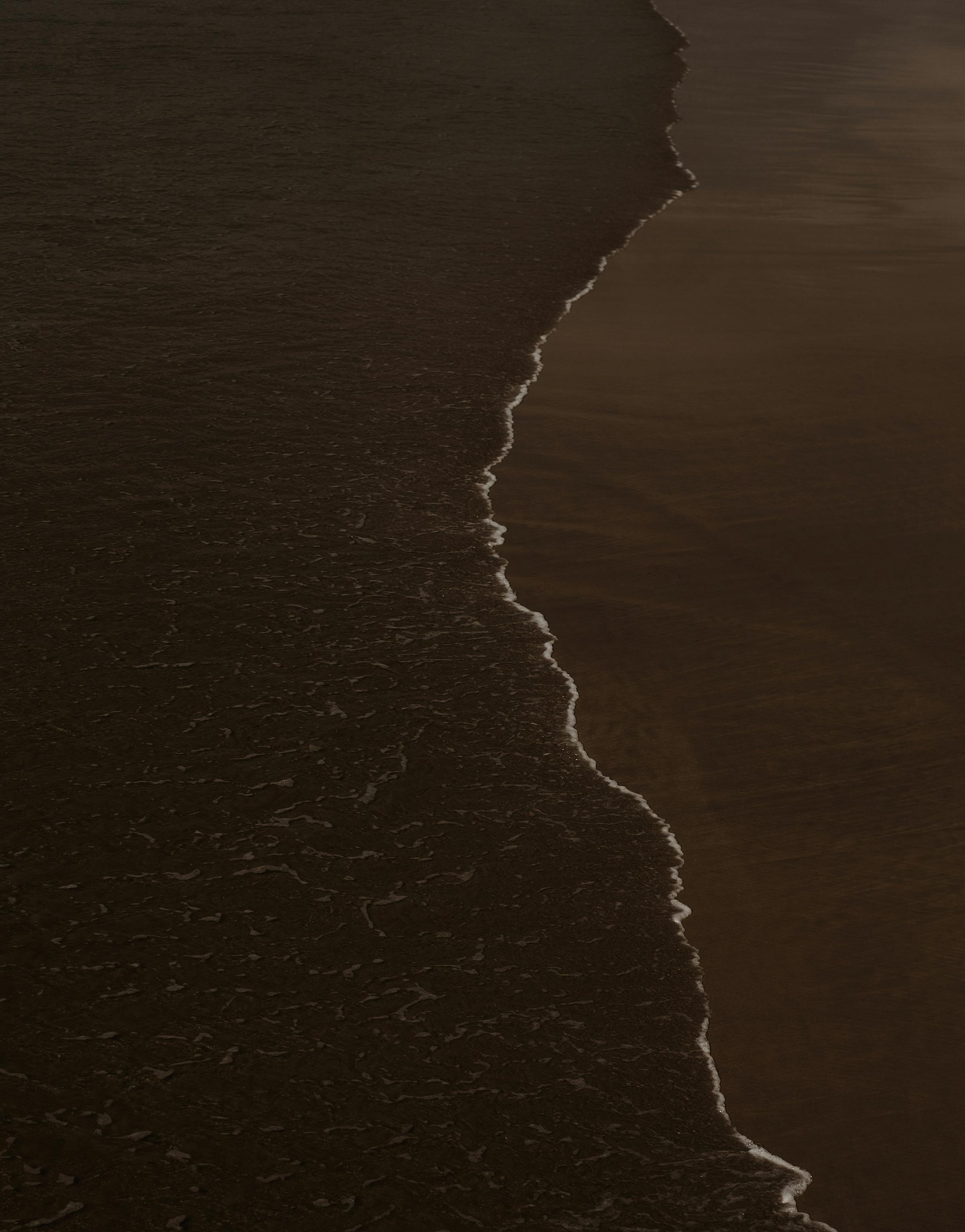A dark photo capturing the contrast between beach water and sand, creating a serene yet moody atmosphere.