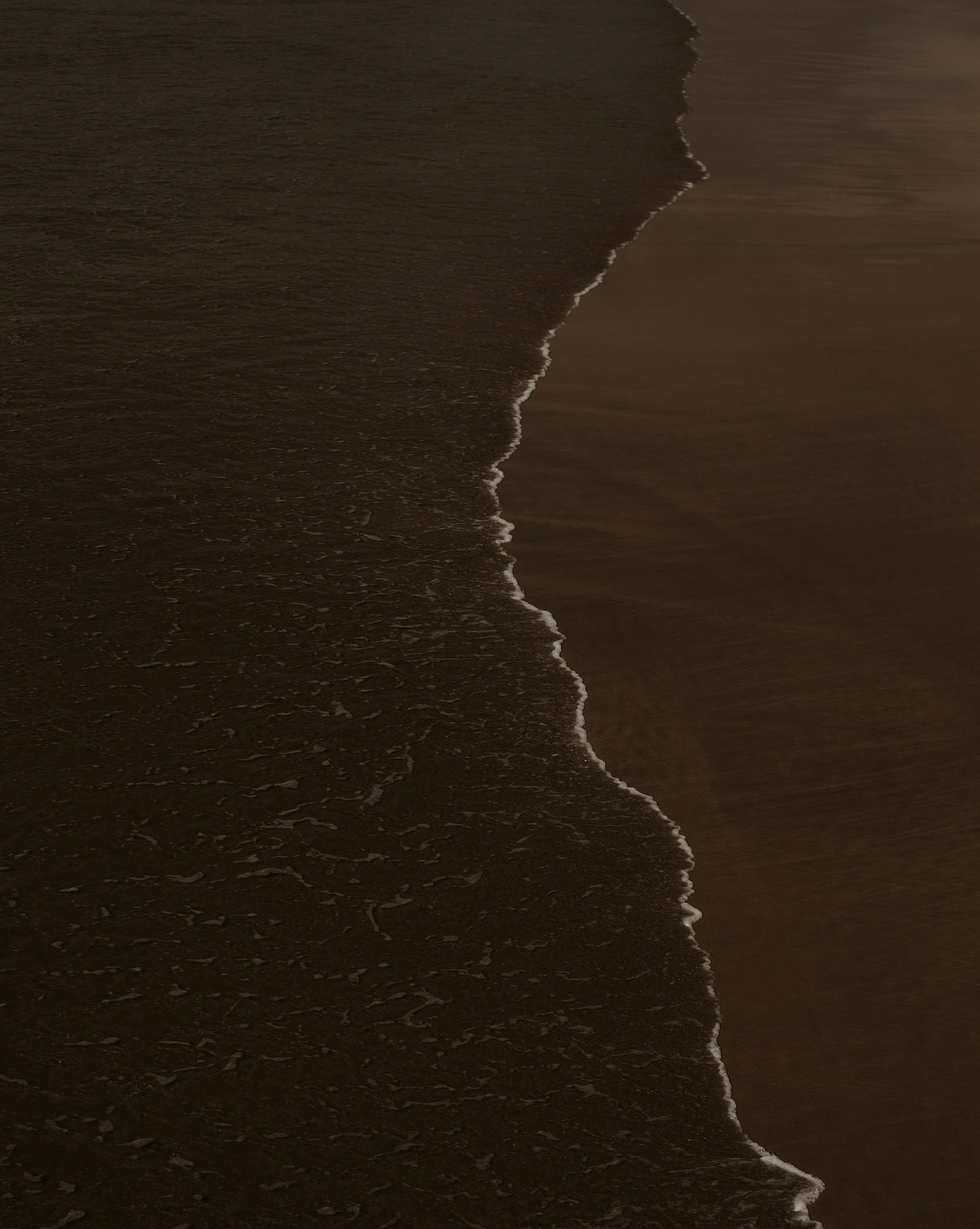A dark photo capturing the contrast between beach water and sand, creating a serene yet moody atmosphere.