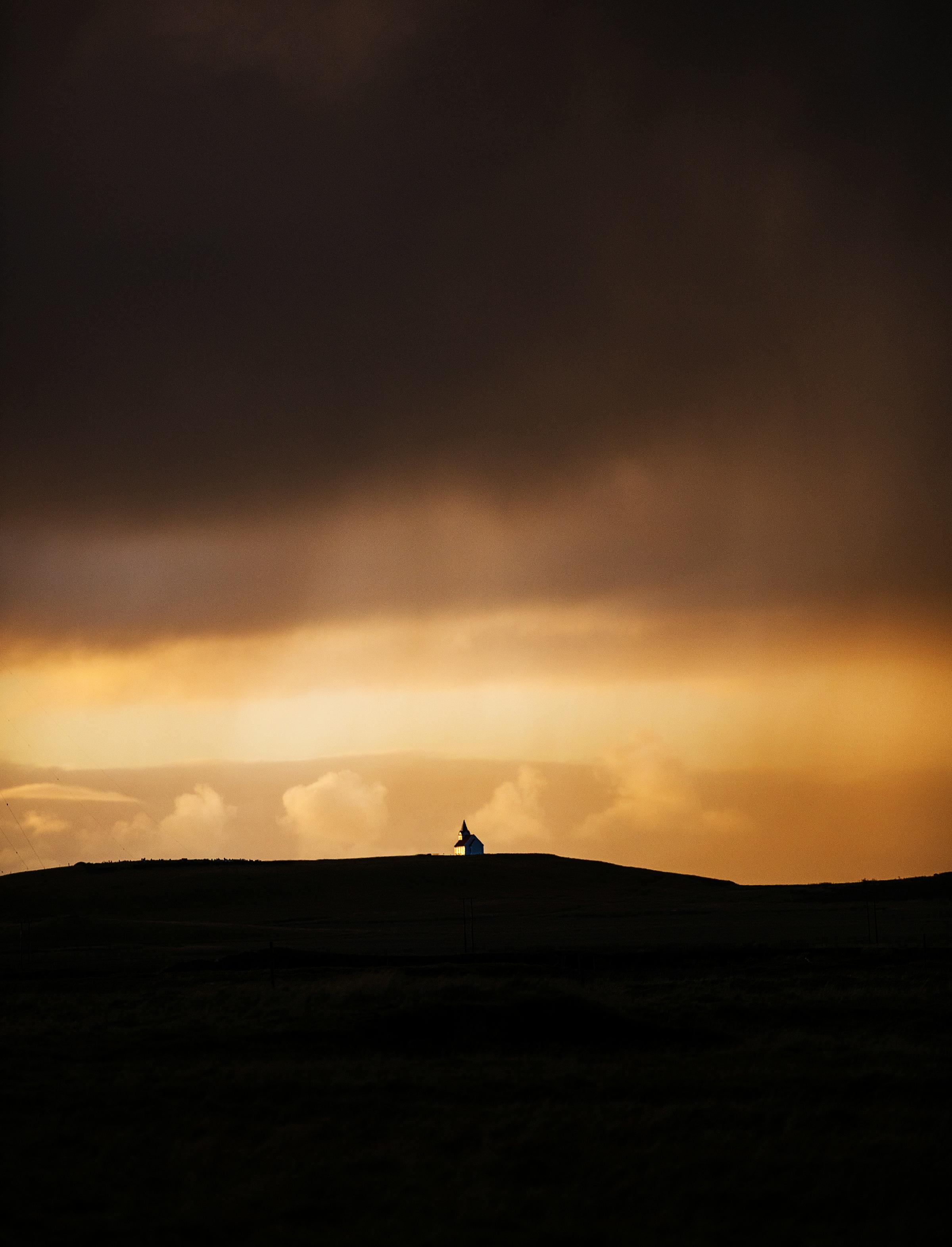 A moody sunset scene with a small, isolated church perched on a hill, illuminated by the warm glow of the setting sun. The expansive sky transitions from golden hues to dark, stormy clouds, creating a dramatic and tranquil atmosphere.