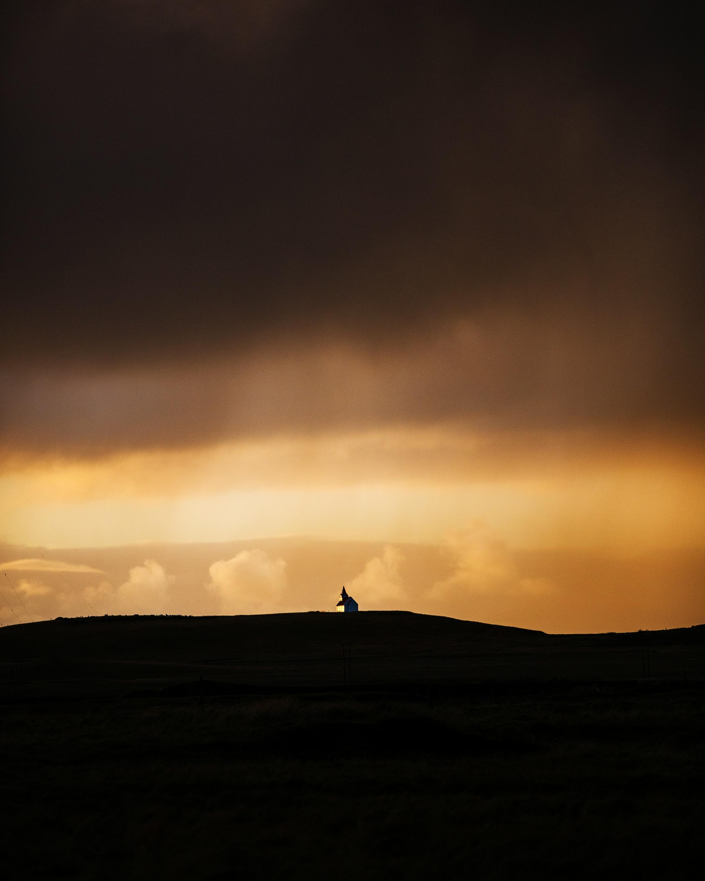 A moody sunset scene with a small, isolated church perched on a hill, illuminated by the warm glow of the setting sun. The expansive sky transitions from golden hues to dark, stormy clouds, creating a dramatic and tranquil atmosphere.