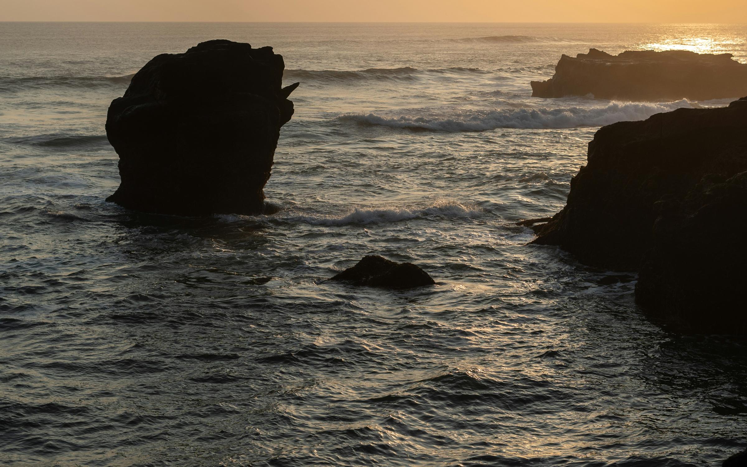 A picturesque sunset over the sea, with vibrant colors reflecting on the water and waves crashing against rocky shores.