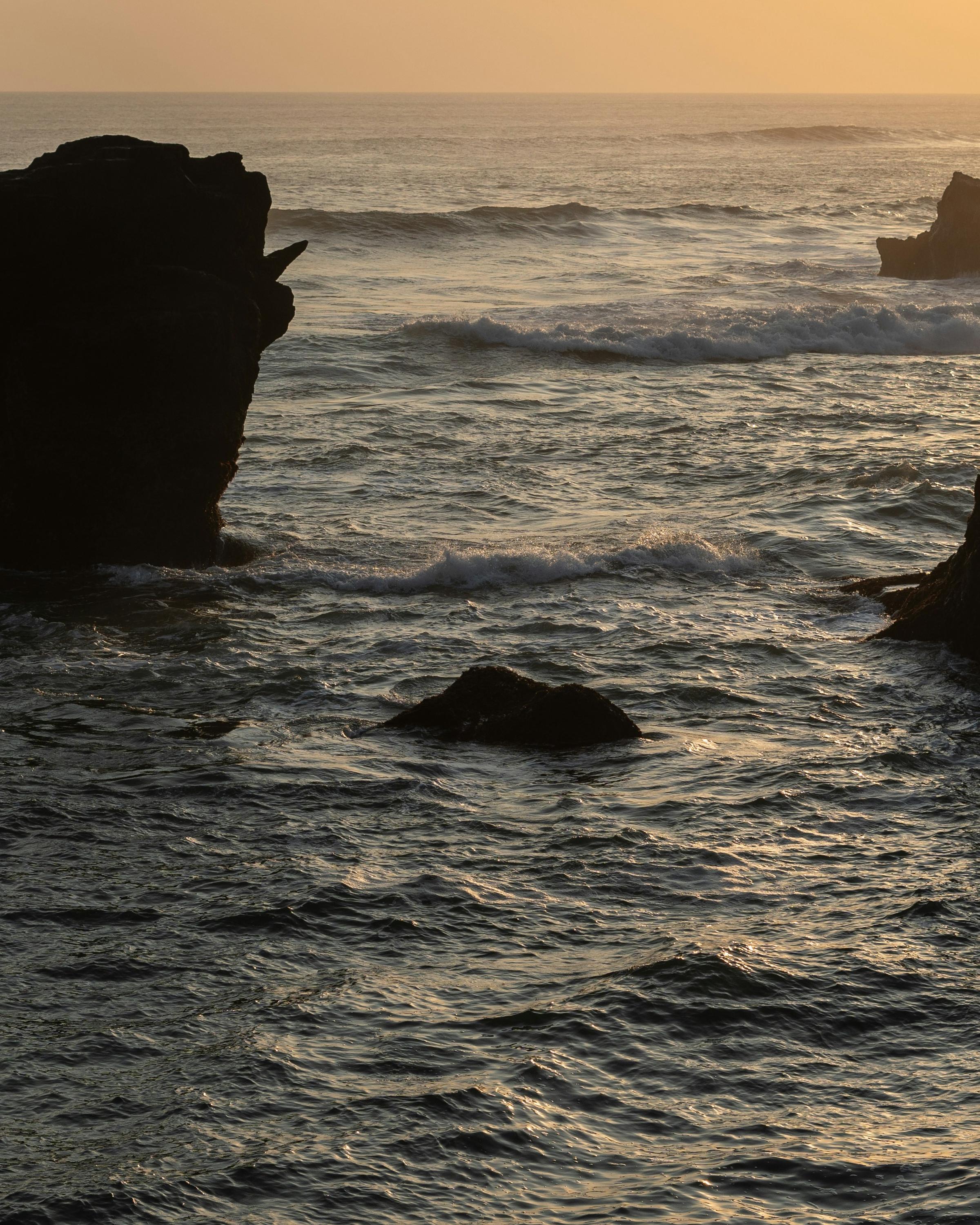 A serene coastal scene at sunset, featuring large, dark rock formations rising from the ocean. The calm waves shimmer in the golden light of the setting sun, creating a tranquil and picturesque atmosphere.
