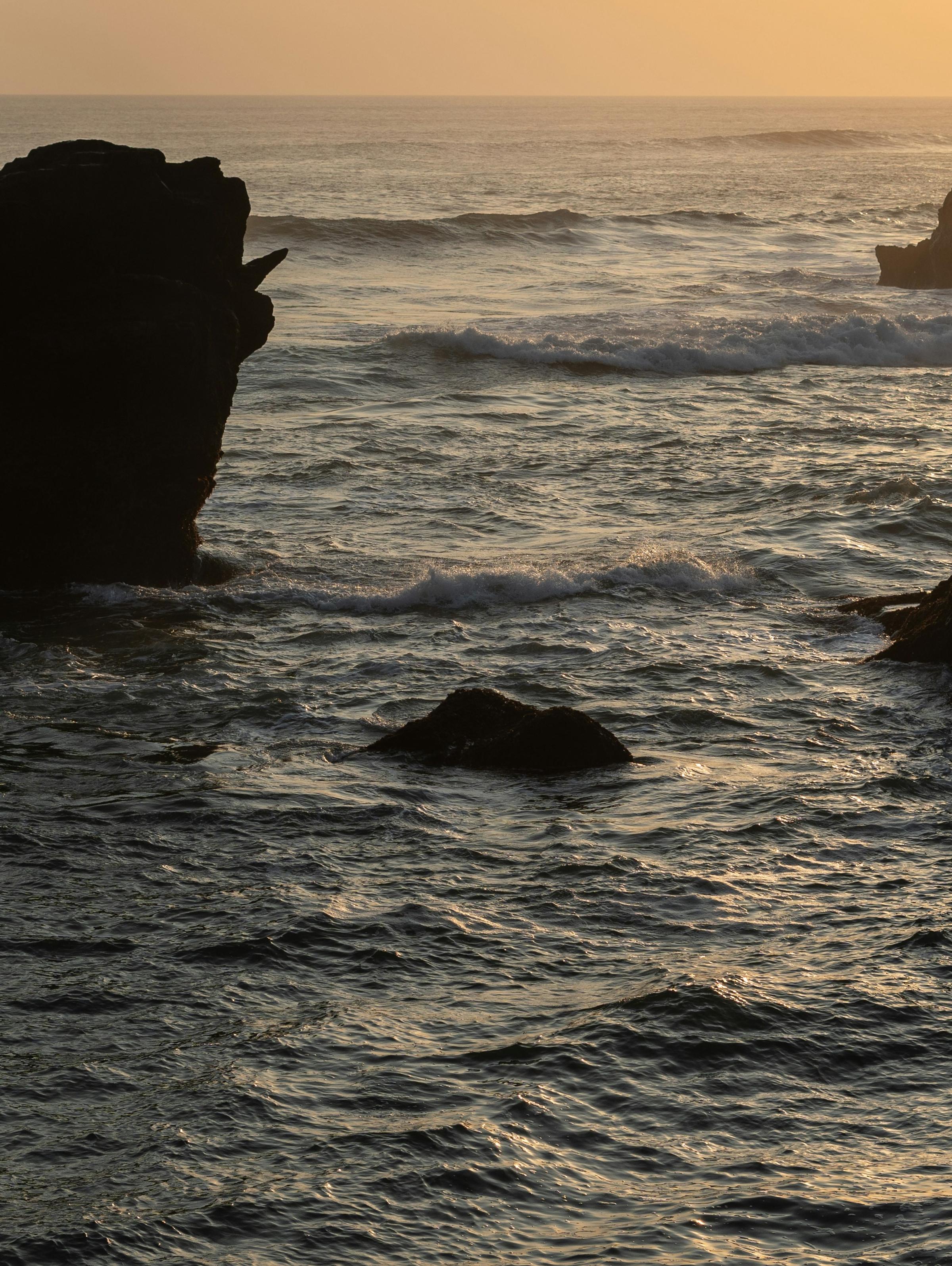 A serene coastal scene at sunset, featuring large, dark rock formations rising from the ocean. The calm waves shimmer in the golden light of the setting sun, creating a tranquil and picturesque atmosphere.