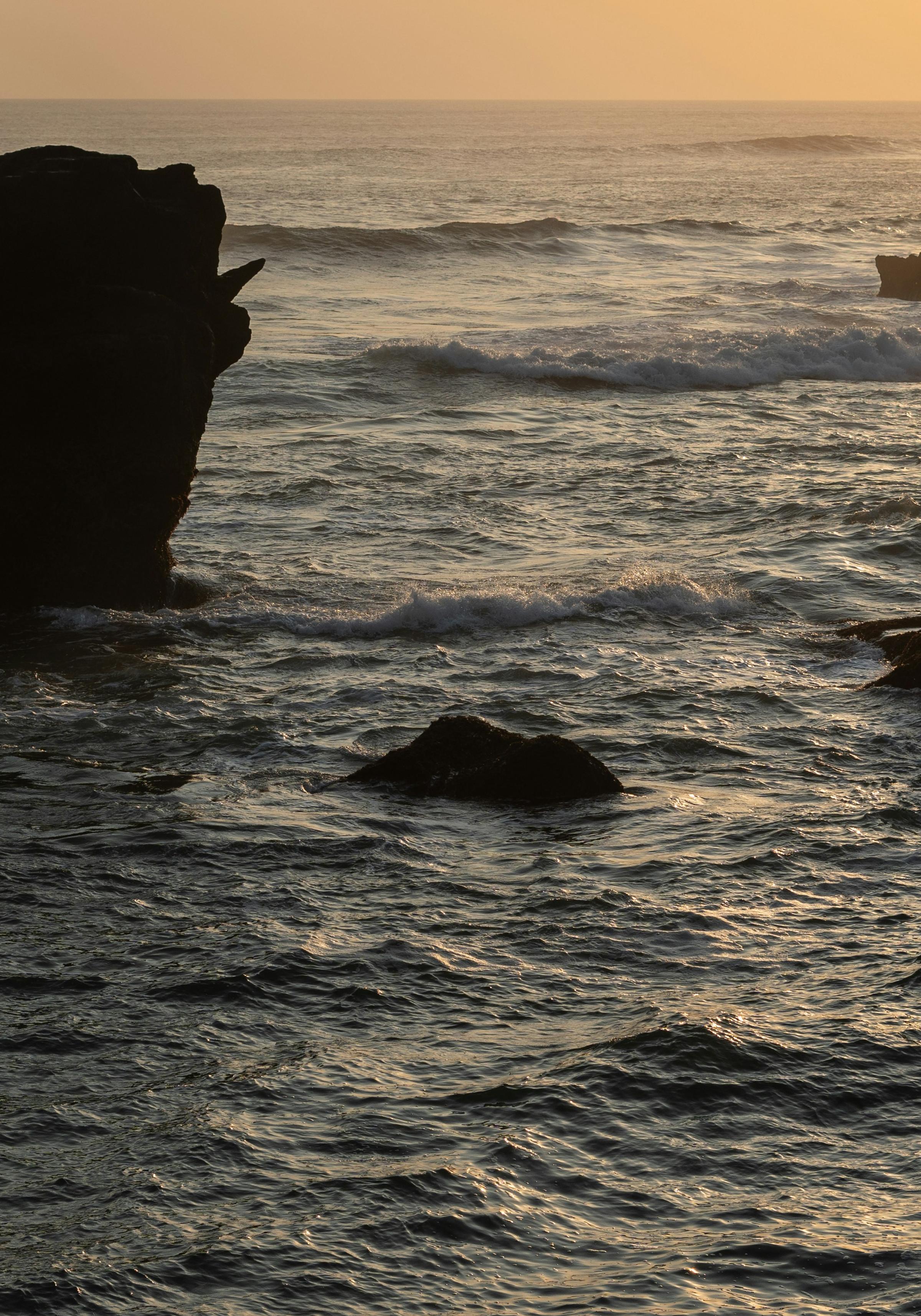 A picturesque sunset over the sea, with vibrant colors reflecting on the water and waves crashing against rocky shores.