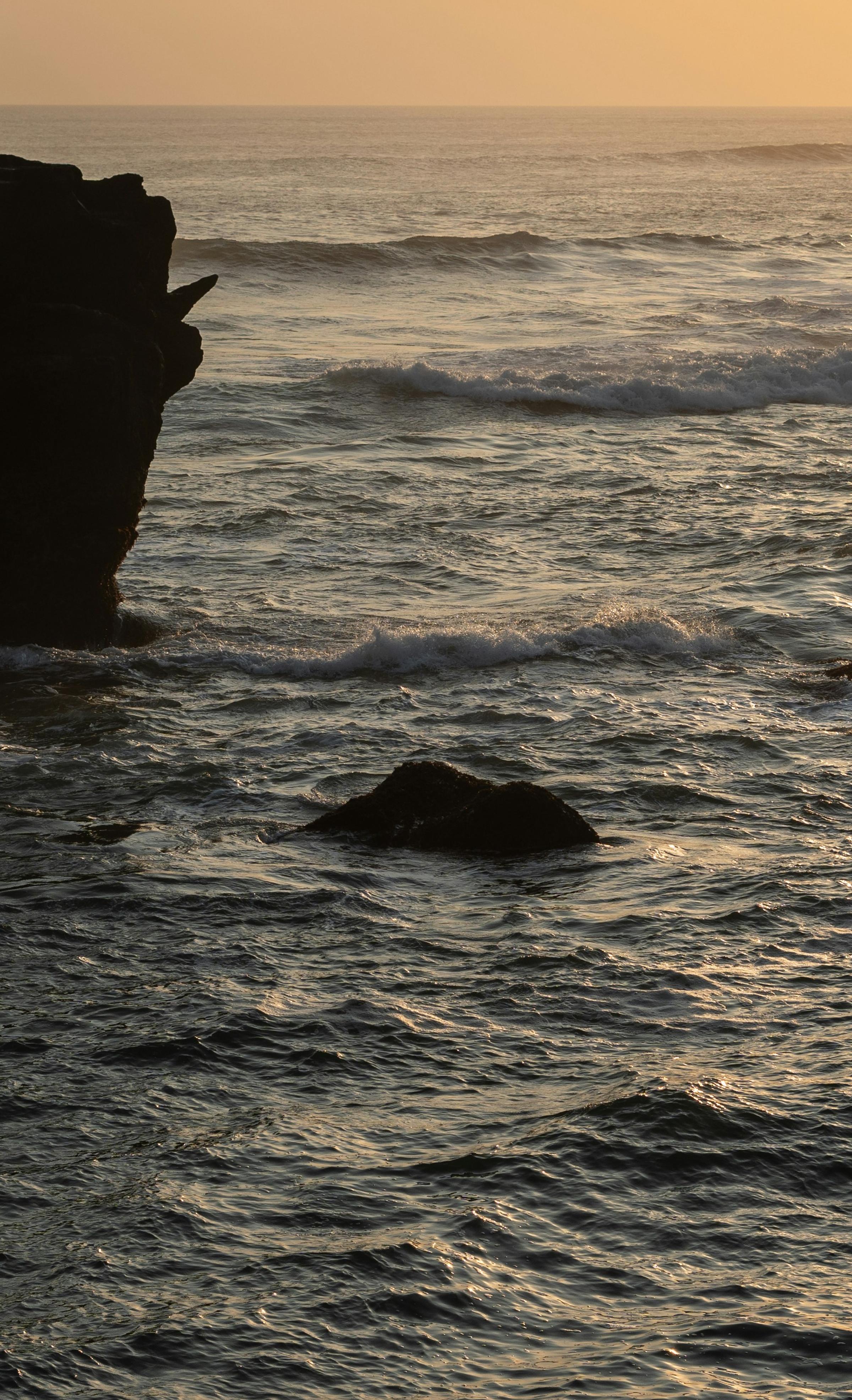 A picturesque sunset over the sea, with vibrant colors reflecting on the water and waves crashing against rocky shores.