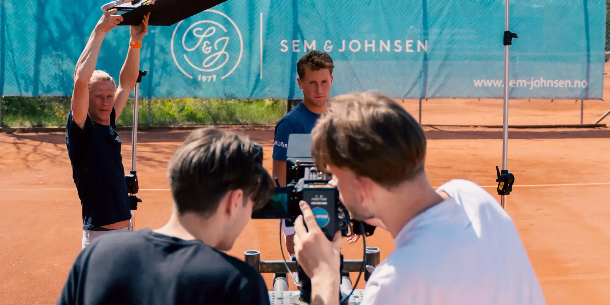 Filmteam med kamera og lysreflektor under en innspilling på en grusbane for tennis