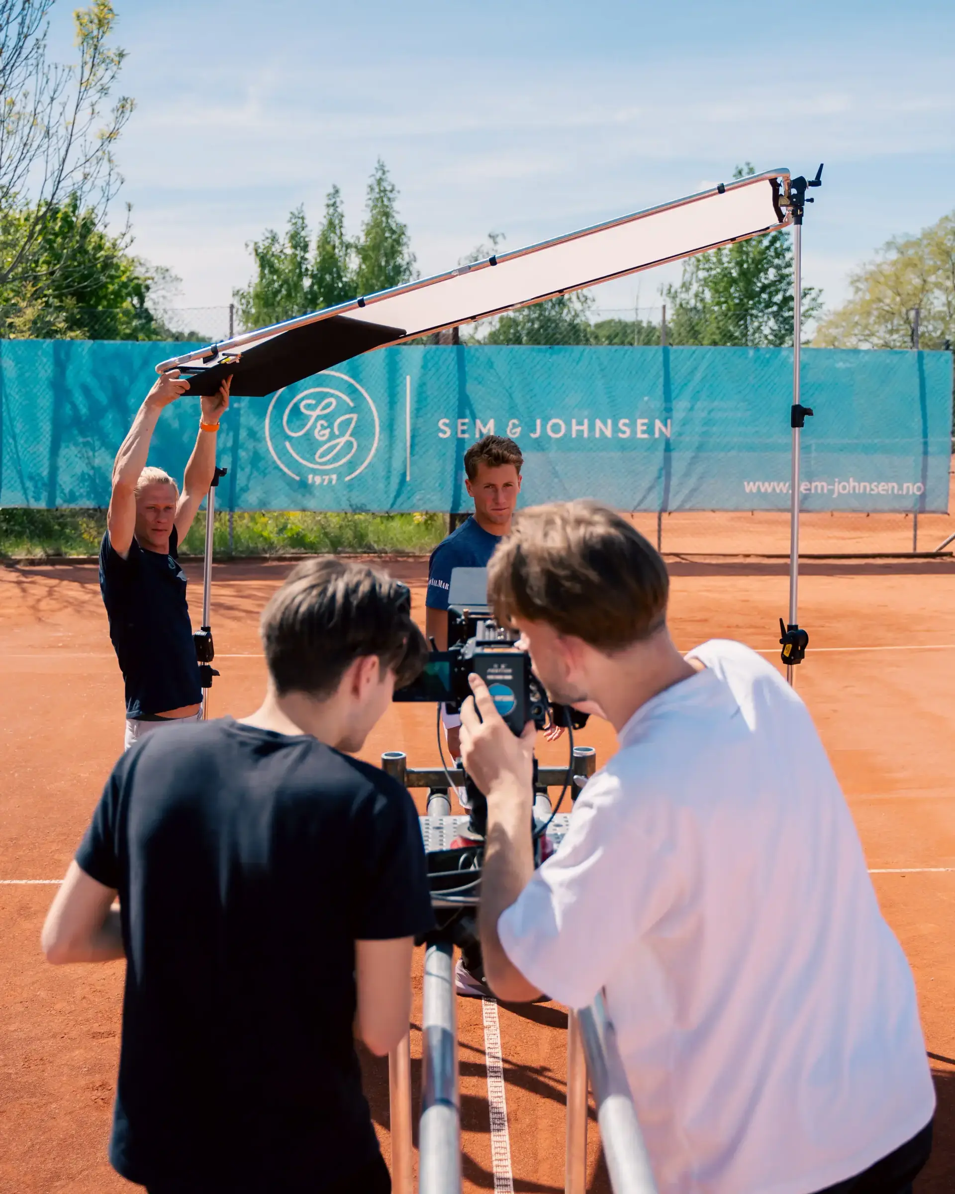 Filmteam med kamera og lysreflektor under en innspilling på en grusbane for tennis