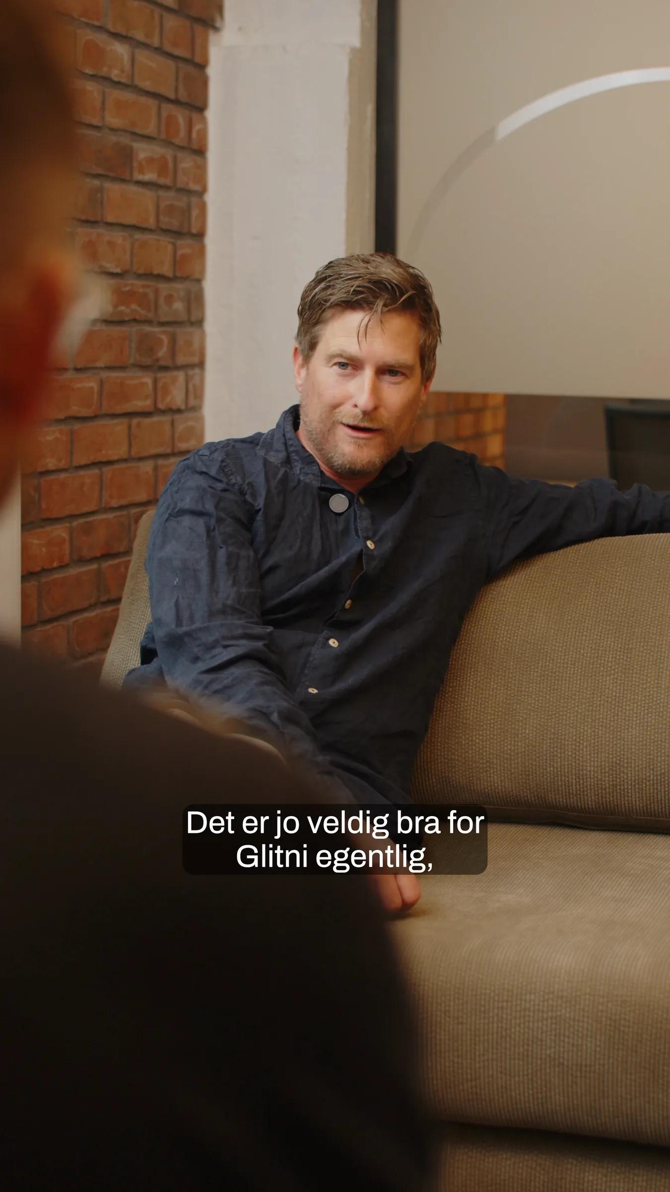 Mann i mørk skjorte sitter i en sofa foran en murvegg og forteller