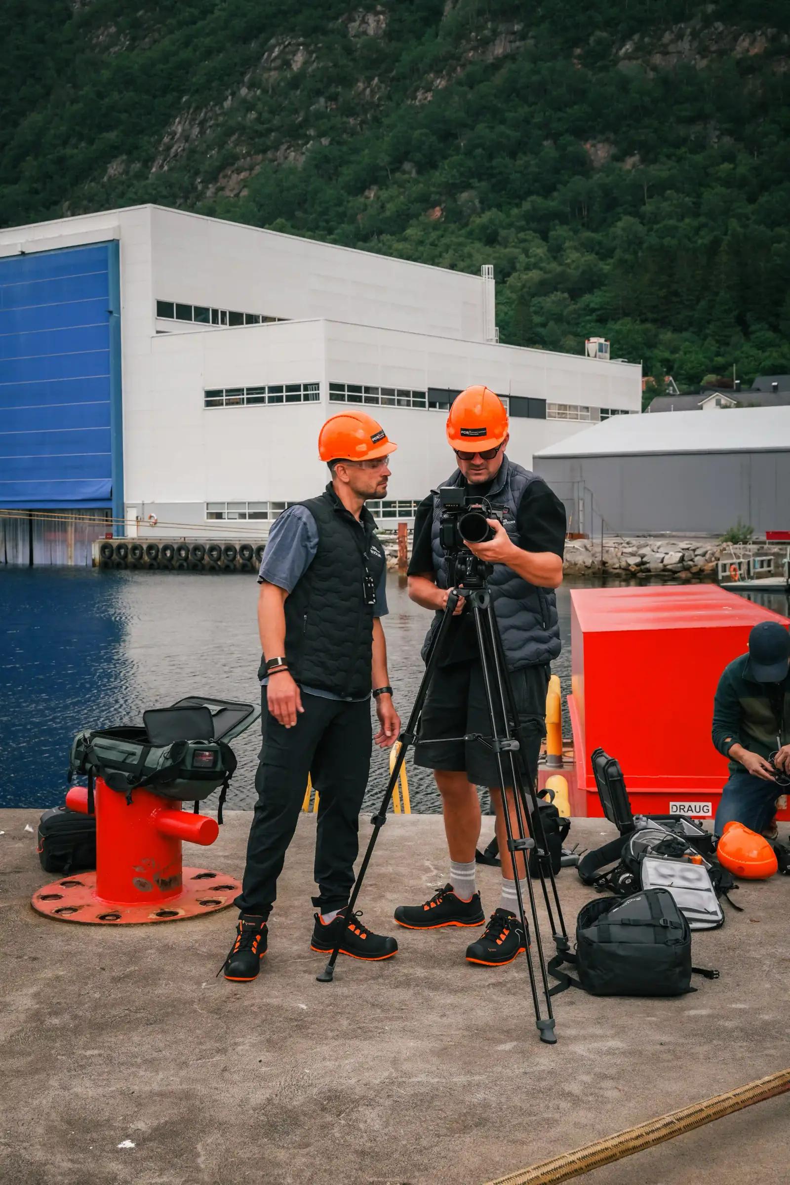 To fotografer med vernehjelmer og kamera på en kai ved et industribygg