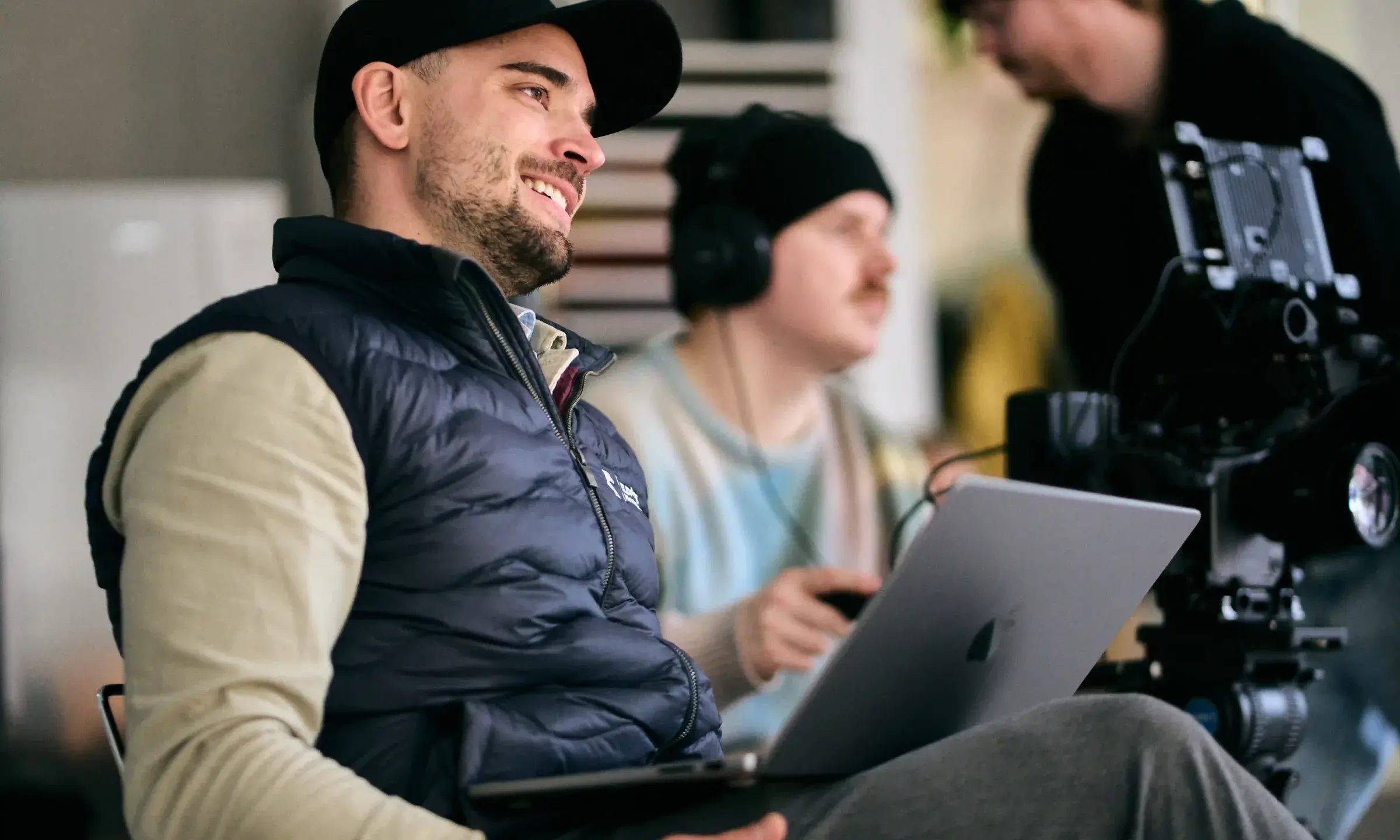 Smilende regissør med laptop på fanget under en innspilling, med kamera og lydtekniker i bakgrunnen