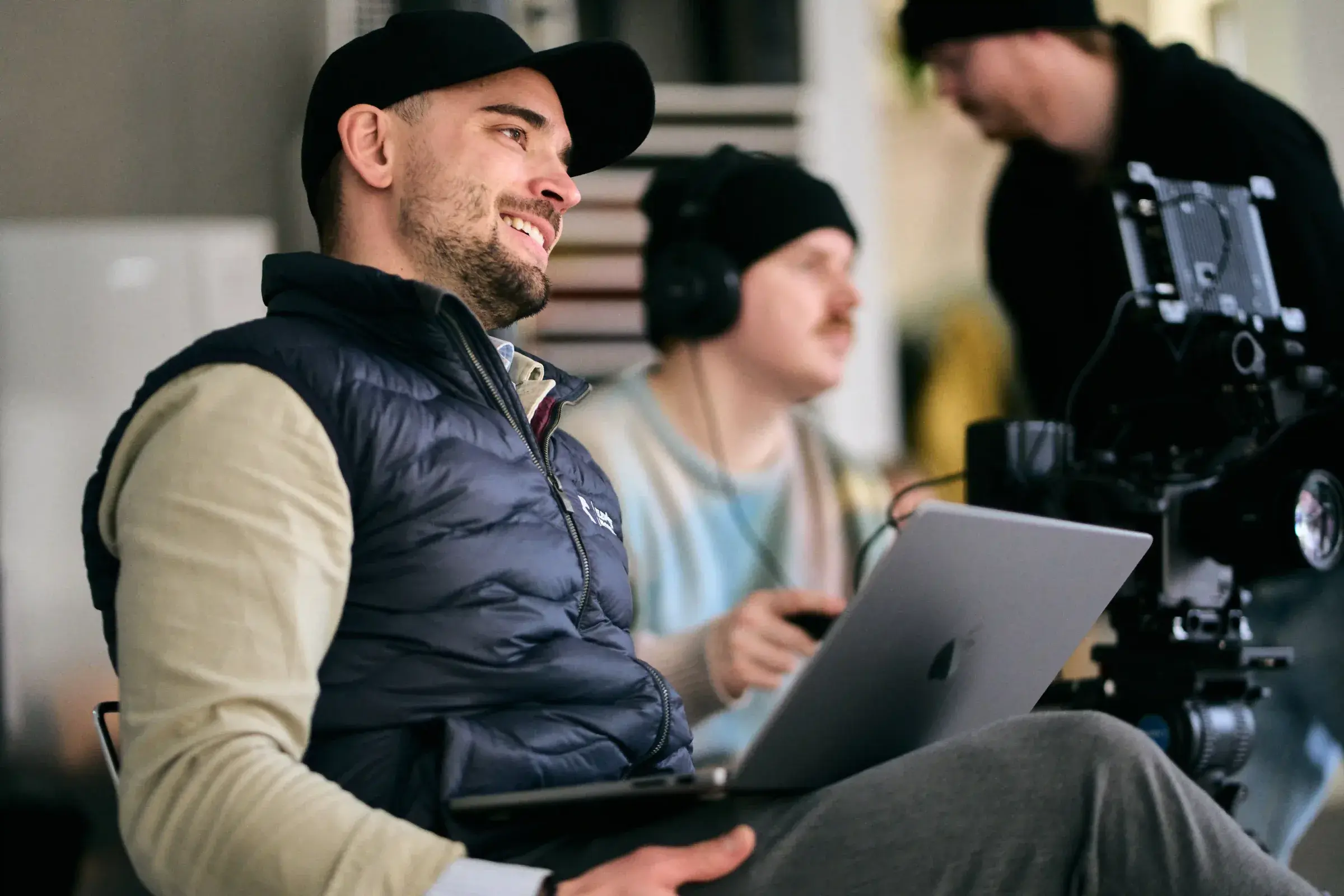 Smilende regissør med laptop på fanget under en innspilling, med kamera og lydtekniker i bakgrunnen