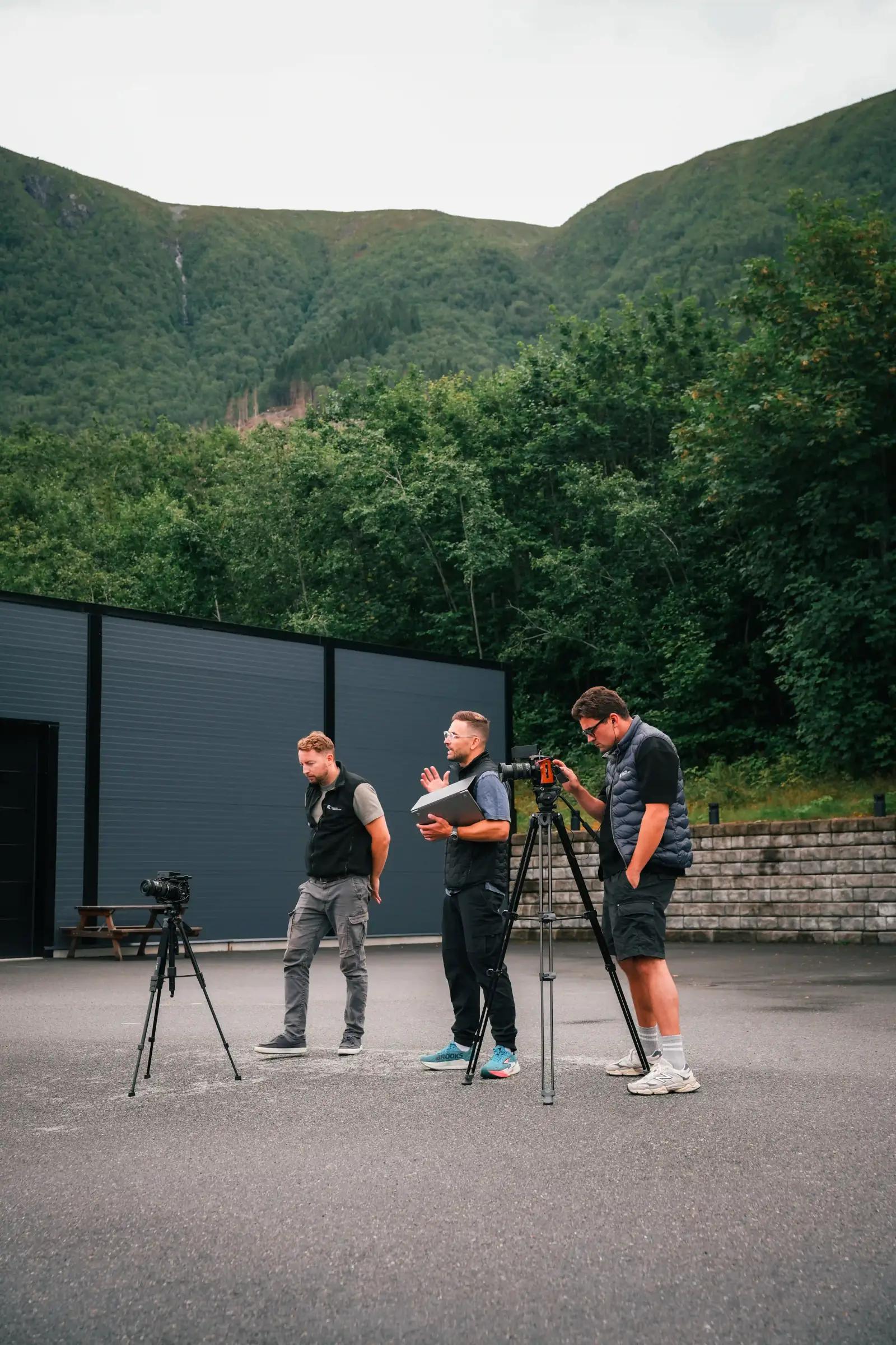 Filmteam med kamera og stativ under et utendørs opptak ved en grønn fjellside