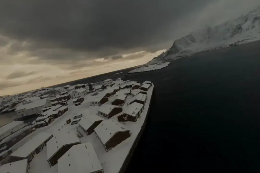 Dronebilde av snødekte rorbuer ved en mørk fjord