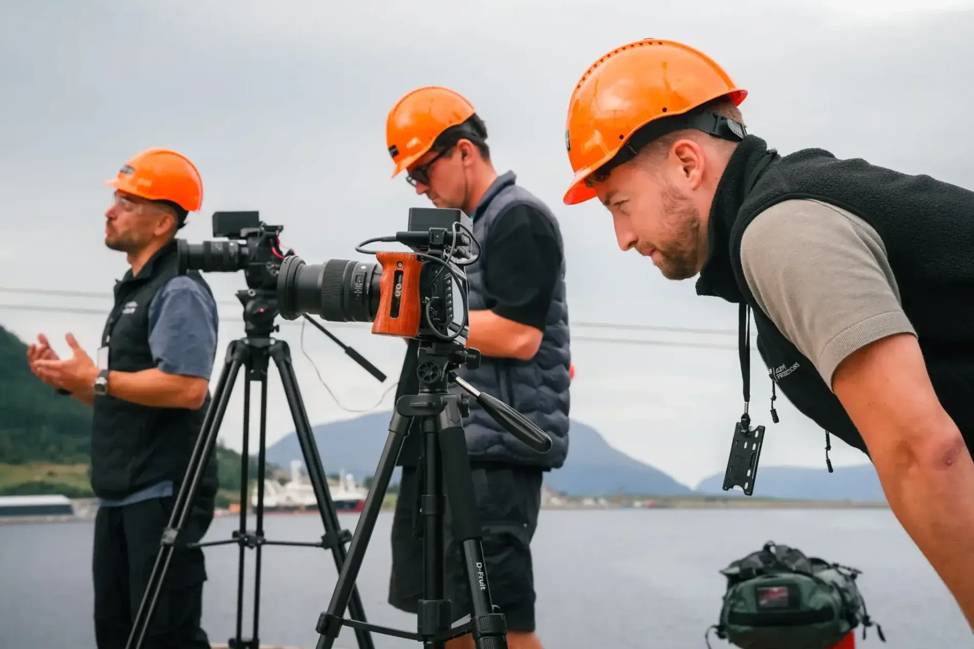 Filmteam med vernehjelmer rigger kamera under et opptak ved sjøen