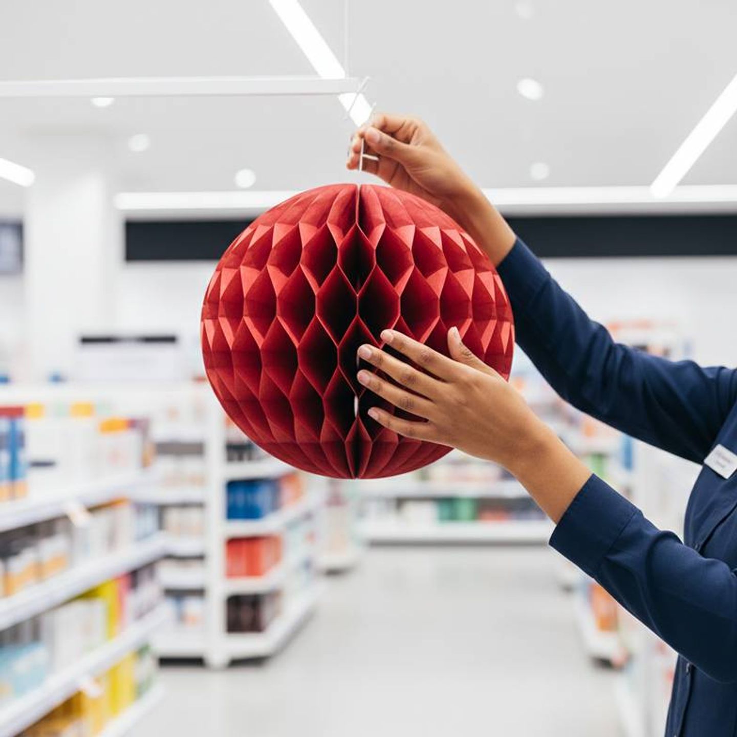A store employee smiling while easily hanging a large red honeycomb paper ornament in a shop window