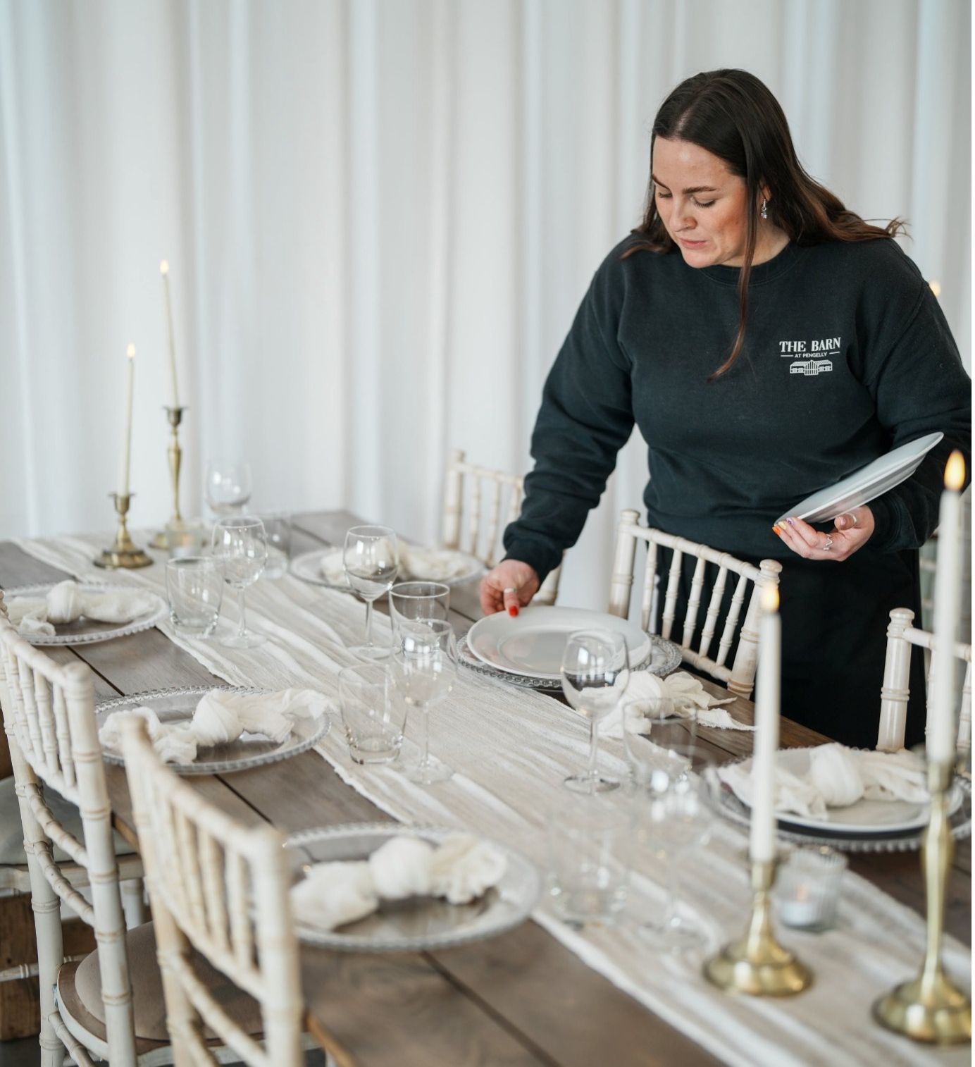Event stylist placing white dinner plates on a rustic wedding reception table with a white runner, brass candlesticks, glassware and Chiavari chairs during venue setup.