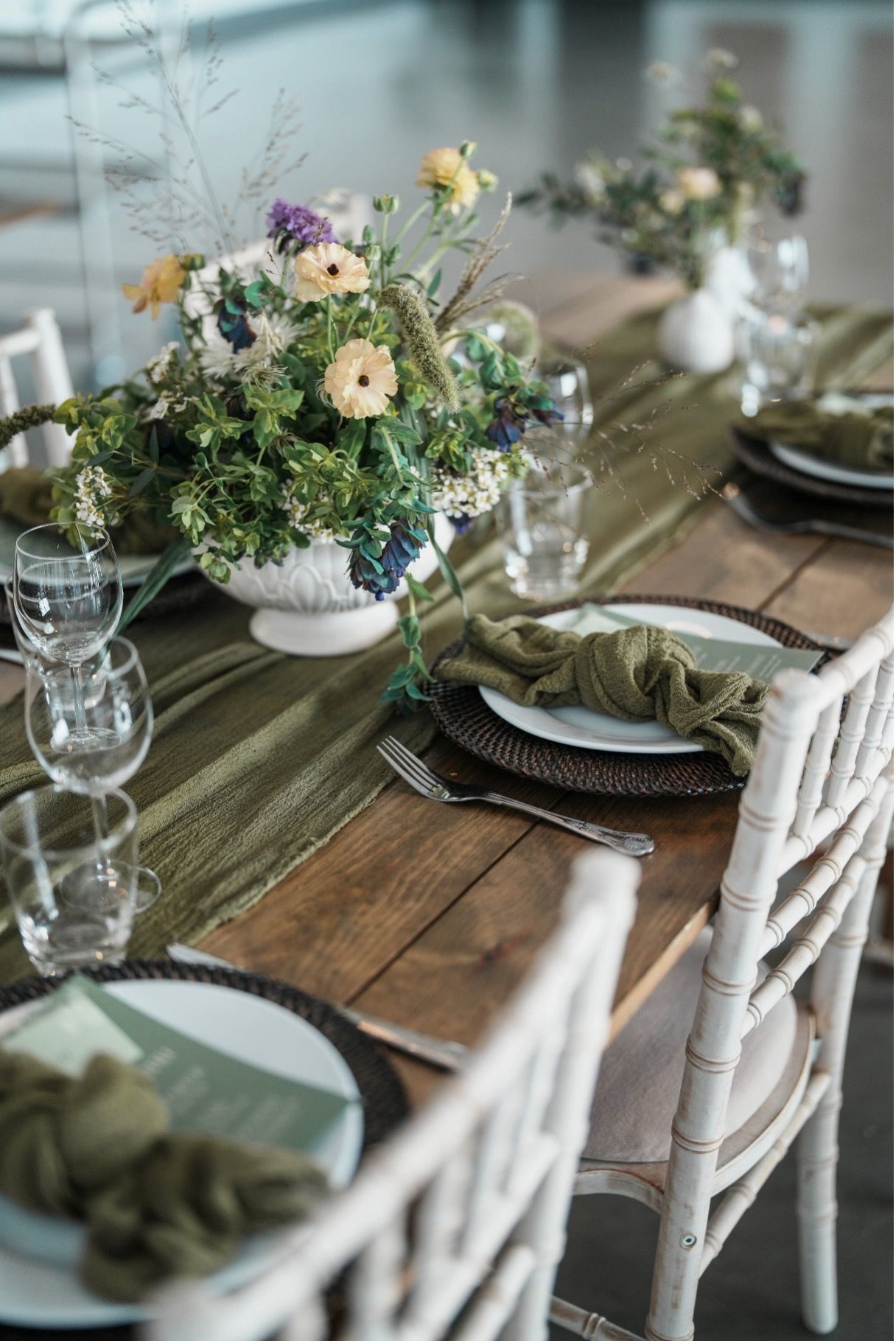 Rustic wooden dining table styled with an olive green gauze runner, lush wildflower centrepieces in white urns, white plates, green knotted napkins and clear glassware for a bohemian wedding reception.