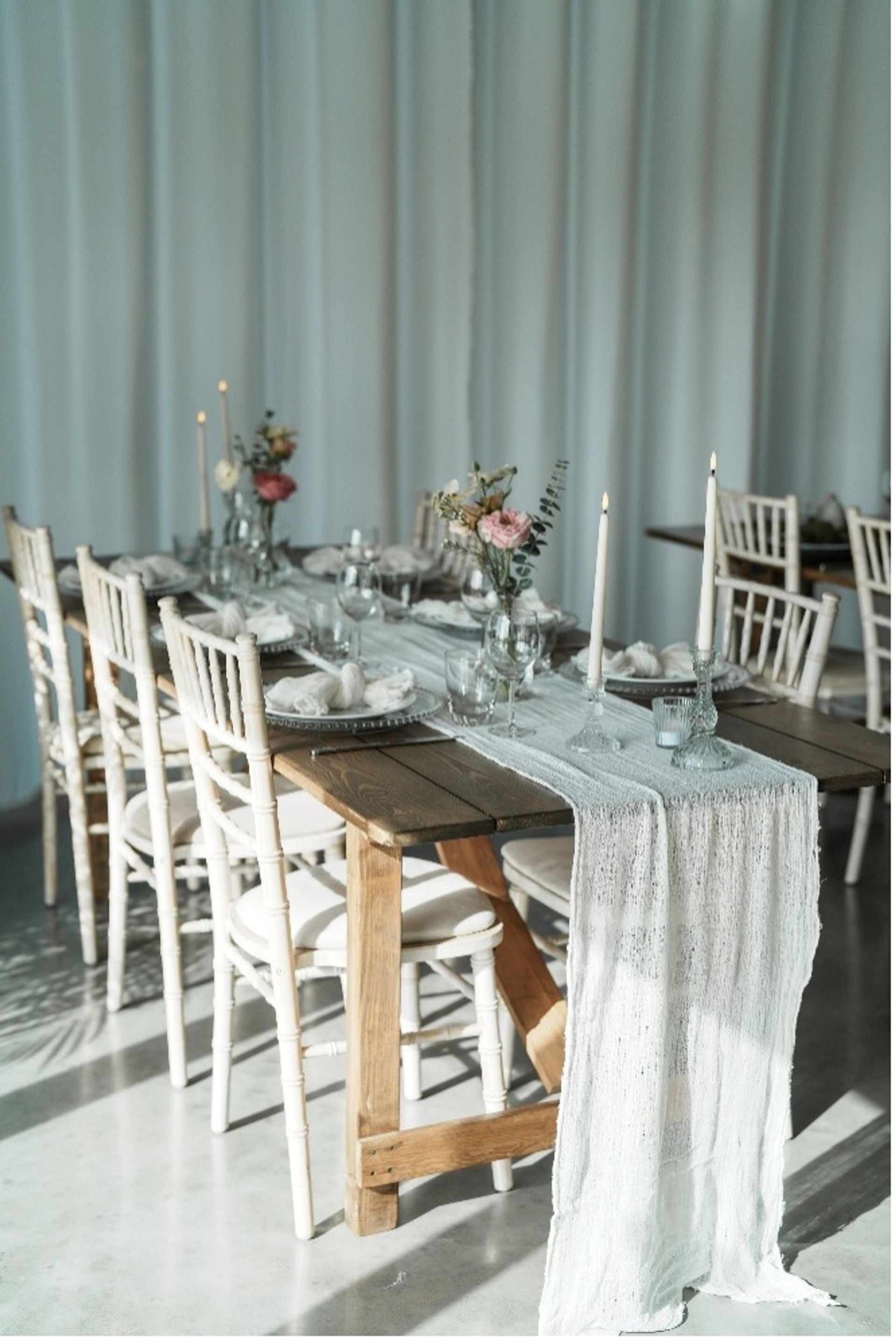 Long wooden banquet table with a white textured runner, brass candlesticks, bud vases of blush flowers, white place settings and Chiavari chairs creating a timeless wedding tablescape.